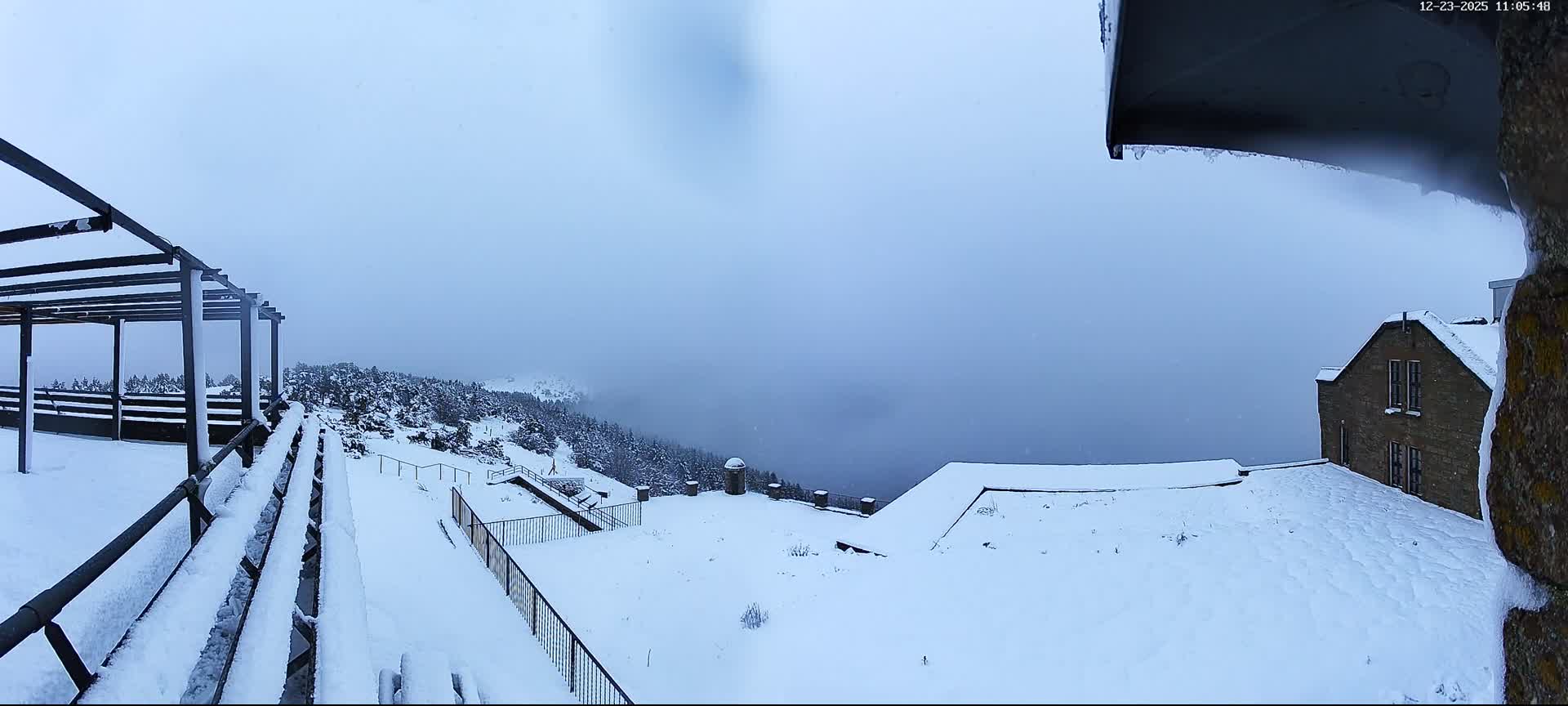A panoramic view reveals a deeply snow-covered mountain landscape with a metal pergola, railings, and evergreen trees, alongside a brick building, all under an overcast sky experiencing light snowfall and significant low visibility from mist obscuring the valley below.