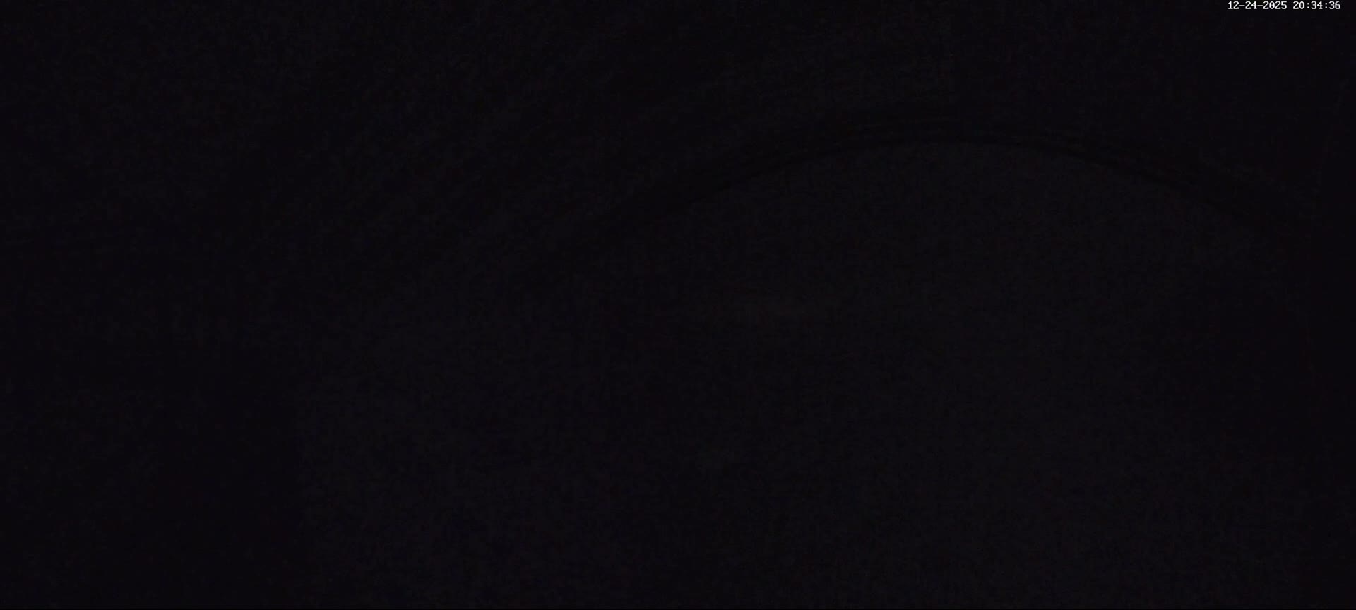 A panoramic view reveals a deeply snow-covered mountain landscape with a metal pergola, railings, and evergreen trees, alongside a brick building, all under an overcast sky experiencing light snowfall and significant low visibility from mist obscuring the valley below.