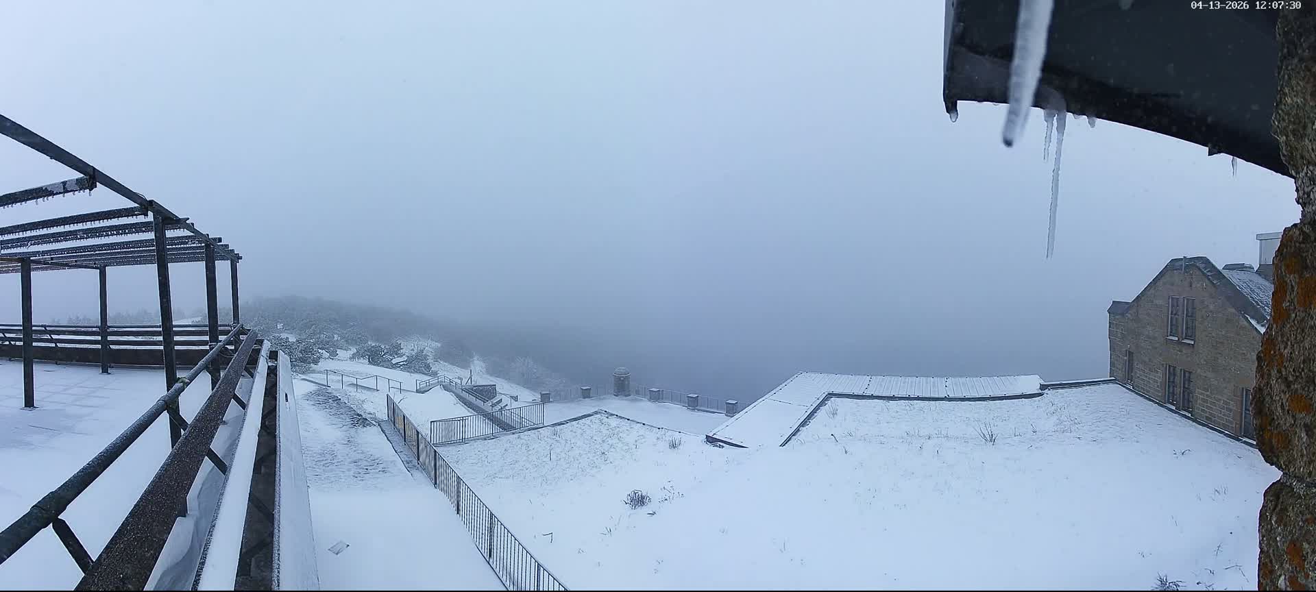 A panoramic view reveals a deeply snow-covered mountain landscape with a metal pergola, railings, and evergreen trees, alongside a brick building, all under an overcast sky experiencing light snowfall and significant low visibility from mist obscuring the valley below.