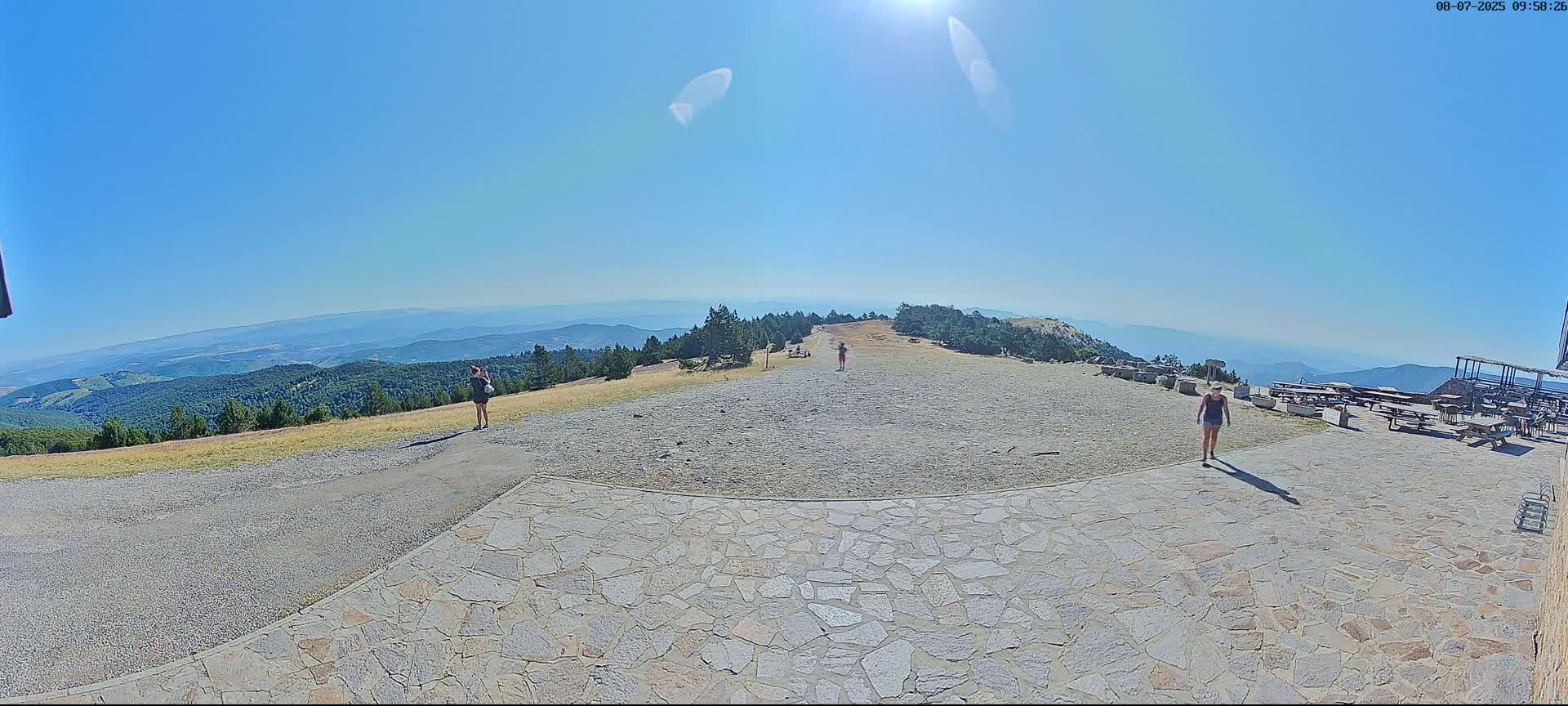 A sunny, clear day reveals a mountaintop vista with several people standing on a stone patio overlooking a vast expanse of forested hills and mountains.