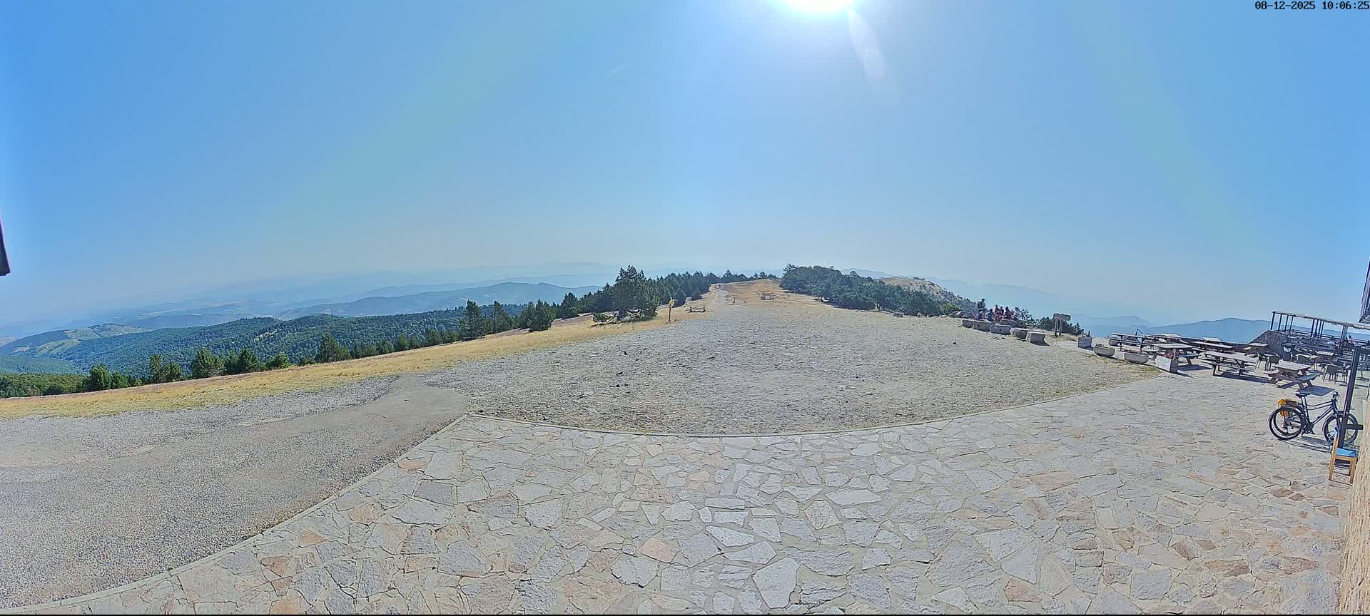 A sunny, clear day reveals a mountaintop vista of rolling hills and forests, with a stone patio area containing picnic tables, a few people, and a bicycle.