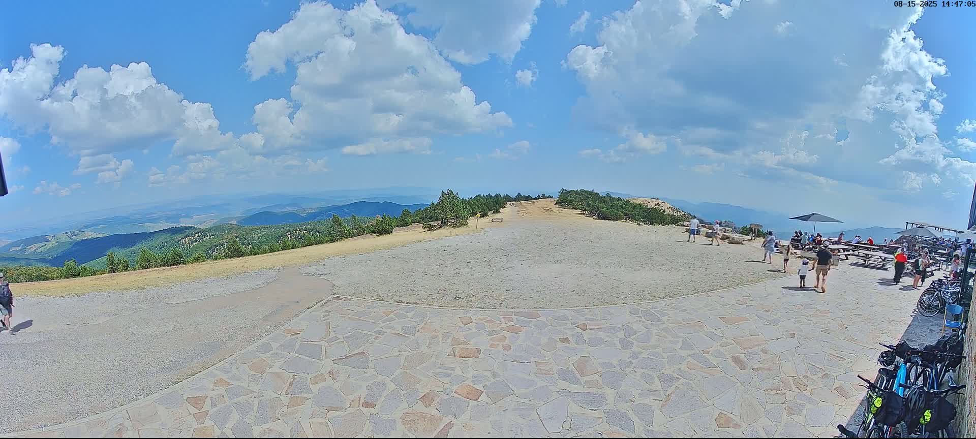 A sunny, clear day reveals a mountaintop vista of rolling hills and forests, with a stone patio area containing picnic tables, a few people, and a bicycle.