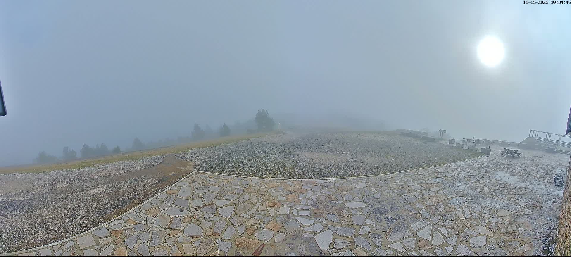 A high-elevation outdoor area with a flagstone patio, gravel ground, and scattered picnic tables is enveloped in dense fog, through which sparse trees and the sun's diffused light are barely visible.
