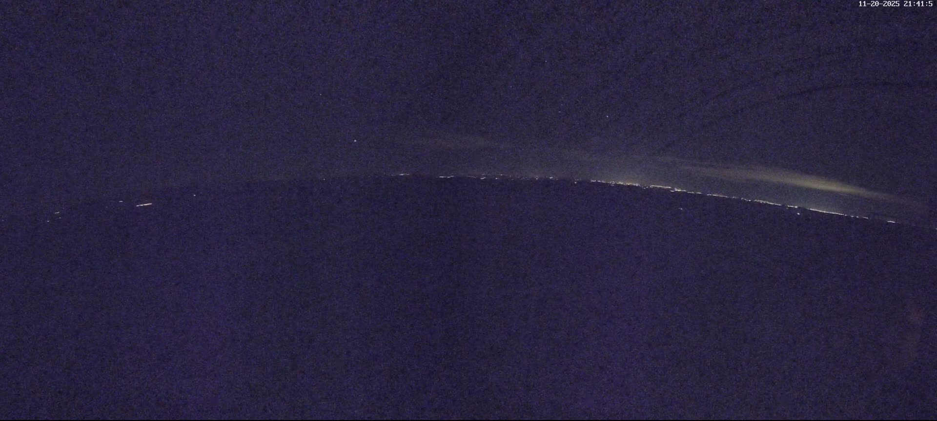 A long band of distant lights from a city or coastline stretches across the horizon under a dark, hazy night sky.