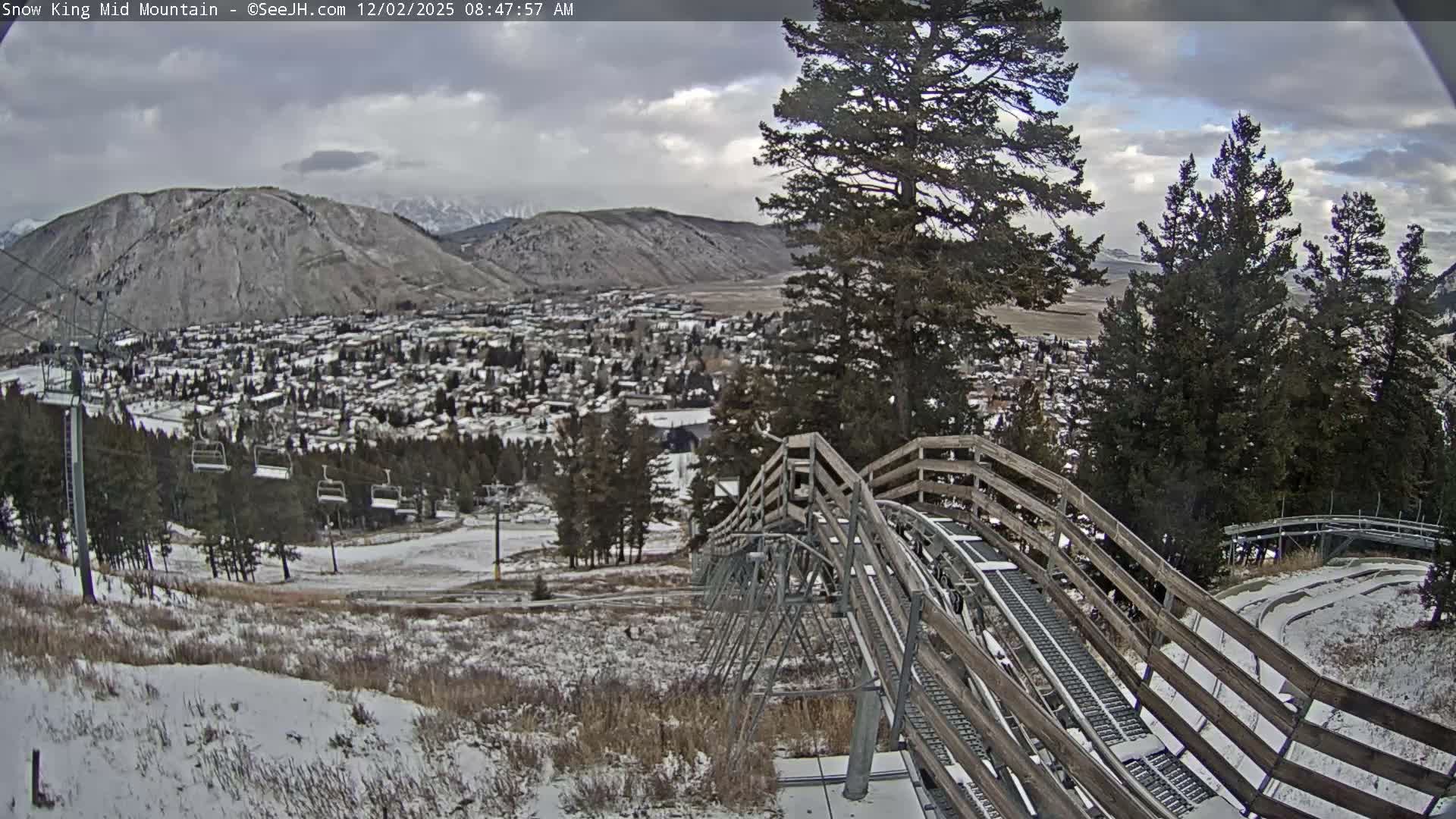A wide shot from a snowy mountain viewpoint reveals an alpine coaster track and ski lift in the foreground, looking down at a snow-dusted town nestled in a valley with large, cloudy mountains in the background under an overcast sky.