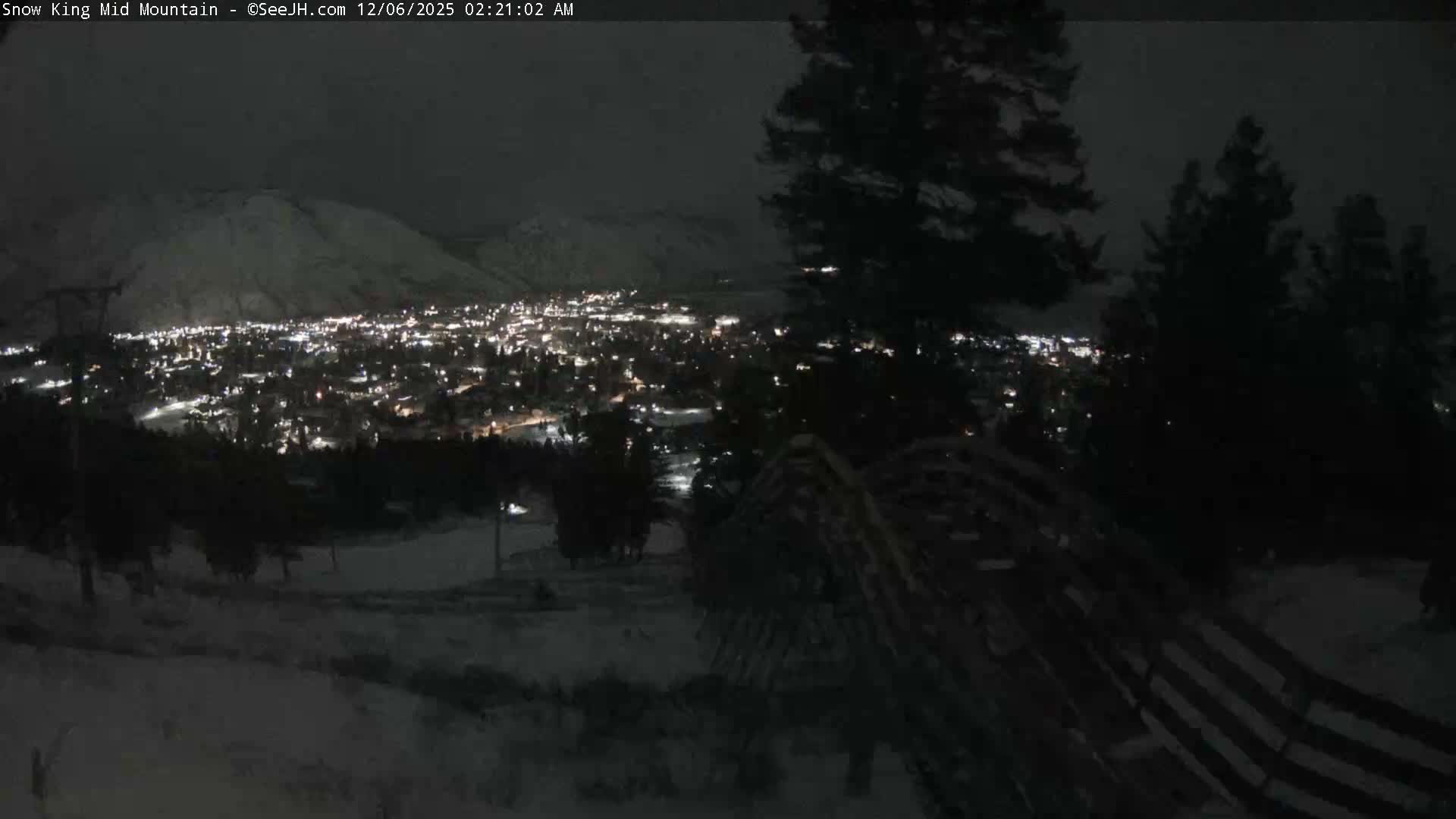 A snow-covered town with twinkling lights lies nestled in a mountain valley at night, framed by dark evergreen trees and a wooden fence in the foreground, all beneath clear, dark skies.