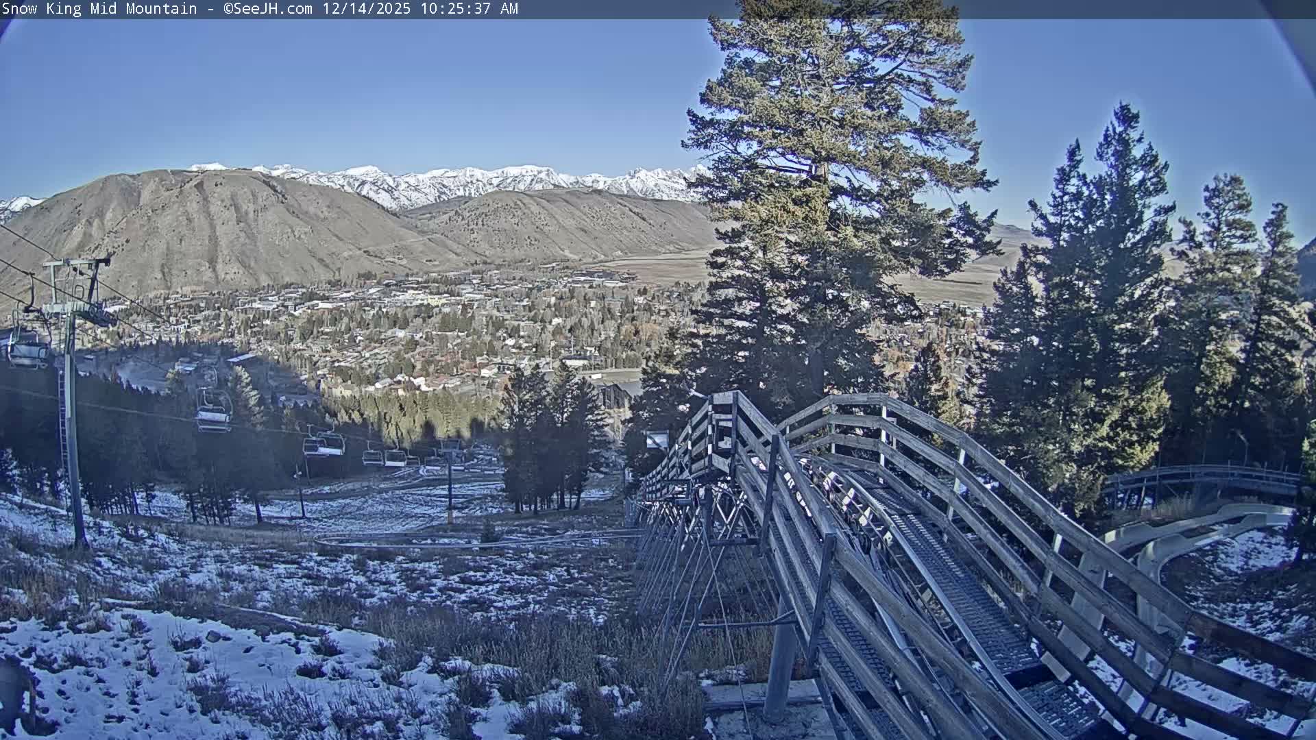 A snow-covered town with twinkling lights lies nestled in a mountain valley at night, framed by dark evergreen trees and a wooden fence in the foreground, all beneath clear, dark skies.