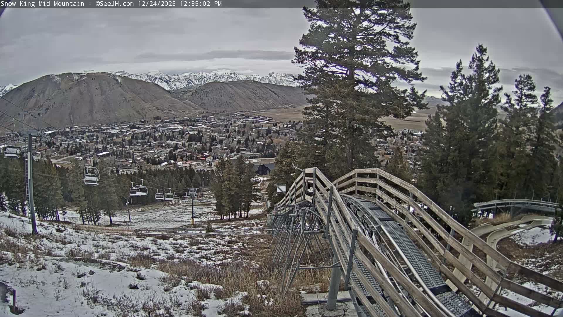 A snow-covered town with twinkling lights lies nestled in a mountain valley at night, framed by dark evergreen trees and a wooden fence in the foreground, all beneath clear, dark skies.