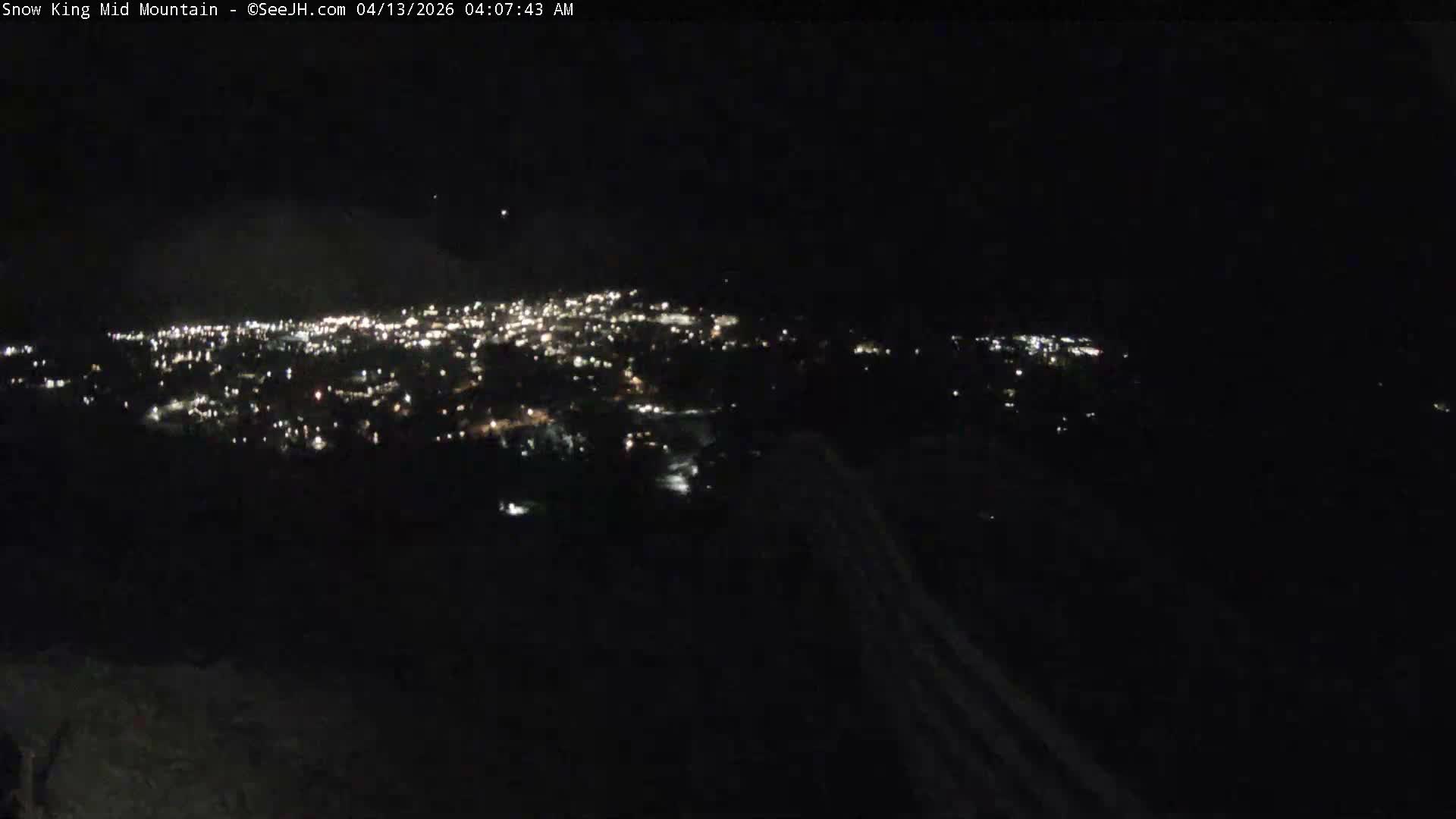 A snow-covered town with twinkling lights lies nestled in a mountain valley at night, framed by dark evergreen trees and a wooden fence in the foreground, all beneath clear, dark skies.