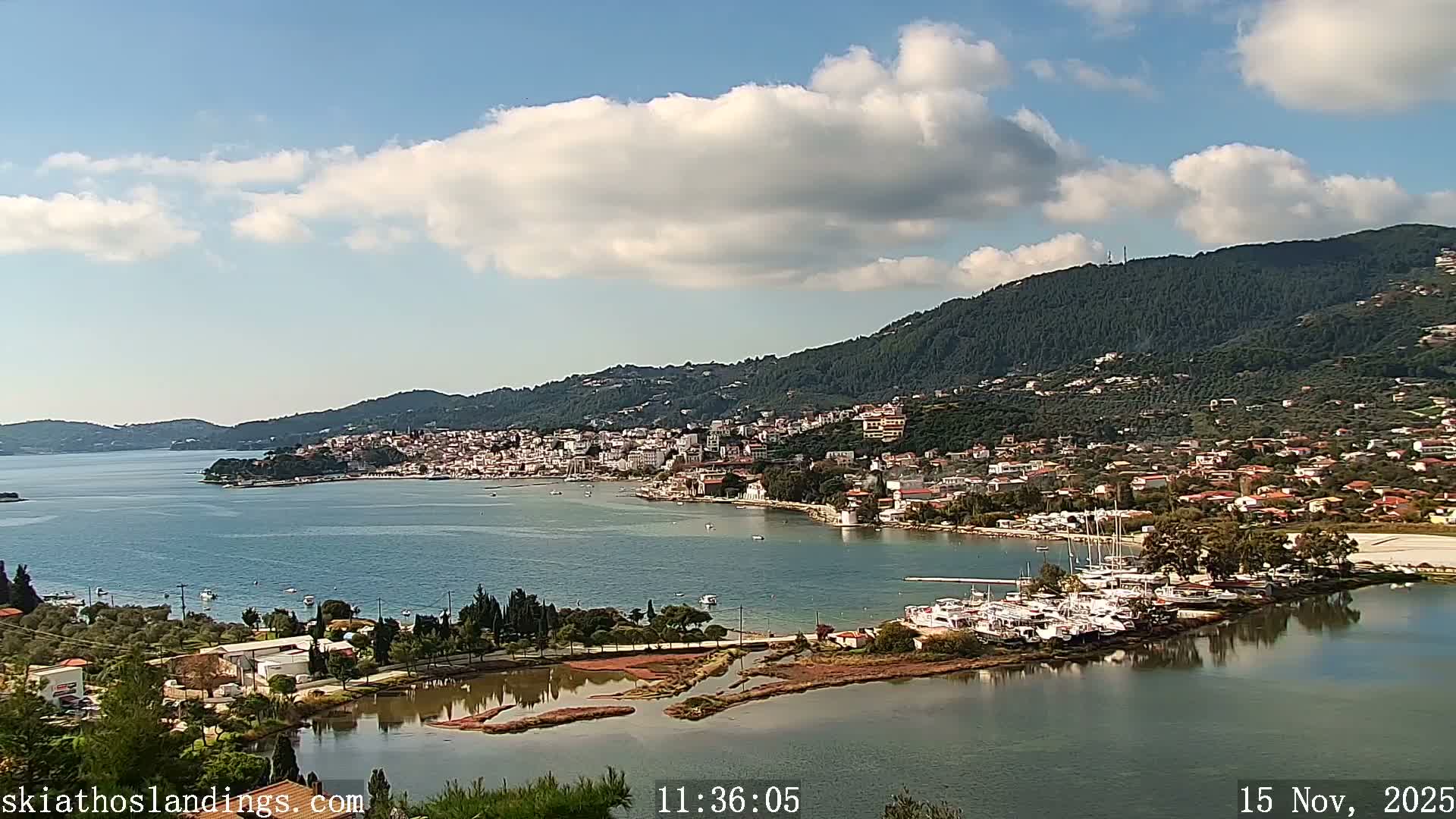 On a clear, sunny day with scattered white clouds, a coastal town featuring white buildings nestled among green, tree-covered hills overlooks a tranquil bay filled with boats, with a smaller, marshy inlet and a marina visible in the foreground.