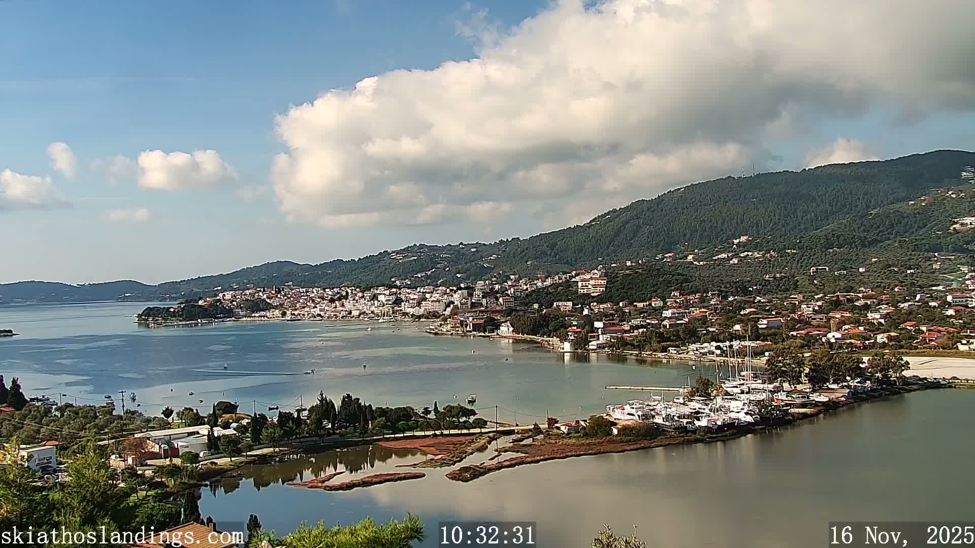 A sunlit panoramic view captures a coastal town sprawling along a bay filled with boats and a marina, backed by densely forested hills under a partly cloudy sky.