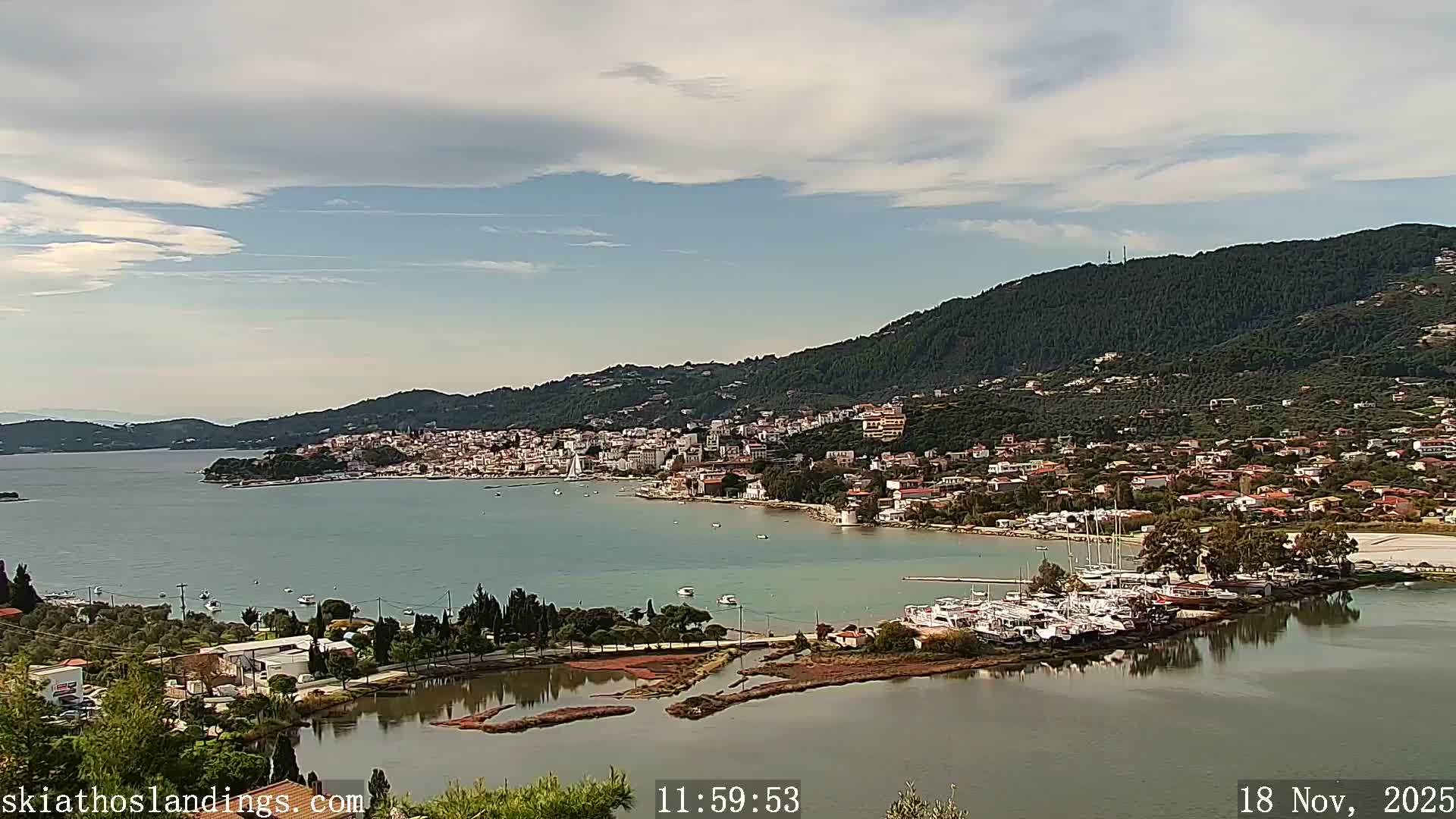 A sunlit panoramic view captures a coastal town sprawling along a bay filled with boats and a marina, backed by densely forested hills under a partly cloudy sky.