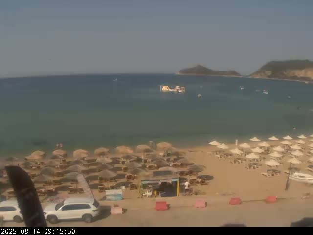 A sandy beach with numerous umbrellas and a calm, clear sea is visible under a sunny sky, with hills in the distance.