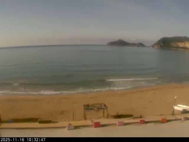 A wide shot reveals a sandy beach with gentle waves meeting the shore, calm blue-grey sea, and several distant, dark landmasses under a clear, bright sky.
