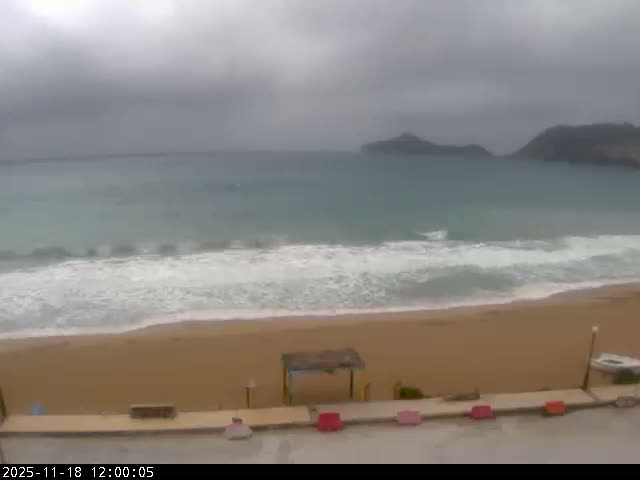 A wide shot reveals a sandy beach with gentle waves meeting the shore, calm blue-grey sea, and several distant, dark landmasses under a clear, bright sky.