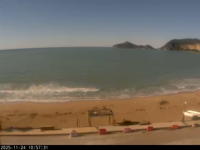 The image captures a sunny day at a sandy beach with gentle waves washing ashore, a vast expanse of greenish-blue sea, and distant hills under a clear blue sky.