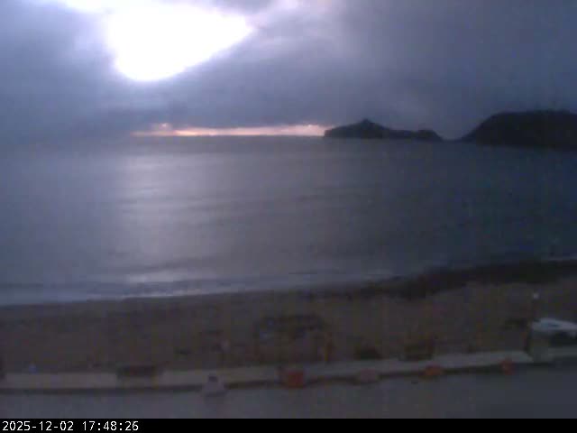 A dusky, overcast coastal scene features a dark sandy beach meeting a calm sea, with distant islands under a heavily clouded sky showing a bright patch of light.