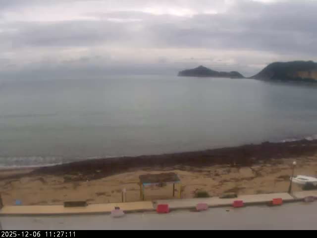 An overcast sky hangs over a coastal scene featuring a grey sea, distant hilly islands, a rocky beach with dark vegetation, and a paved promenade with red barriers in the foreground.