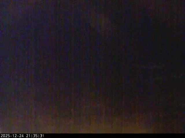 Under a heavily overcast sky, waves crash onto a sandy beach with a few structures, stretching towards distant hazy islands.