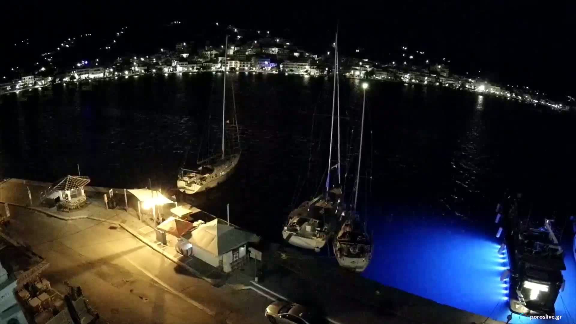 At night, several sailboats are docked in a calm harbor next to a town with lights illuminating the buildings and streets where cars are driving.