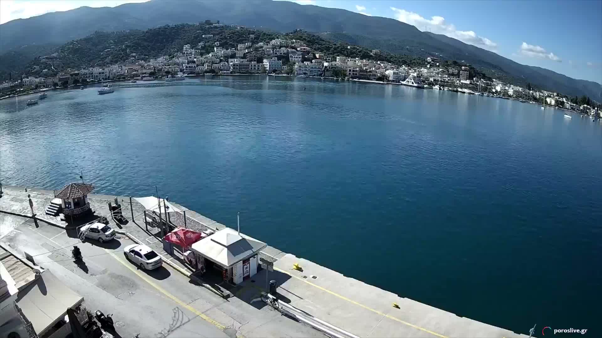 An elevated view captures a picturesque coastal town with white buildings ascending green mountains, overlooking a calm blue bay dotted with boats, all under a bright, partly cloudy sky, with a concrete pier featuring small structures, cars, and people in the foreground.