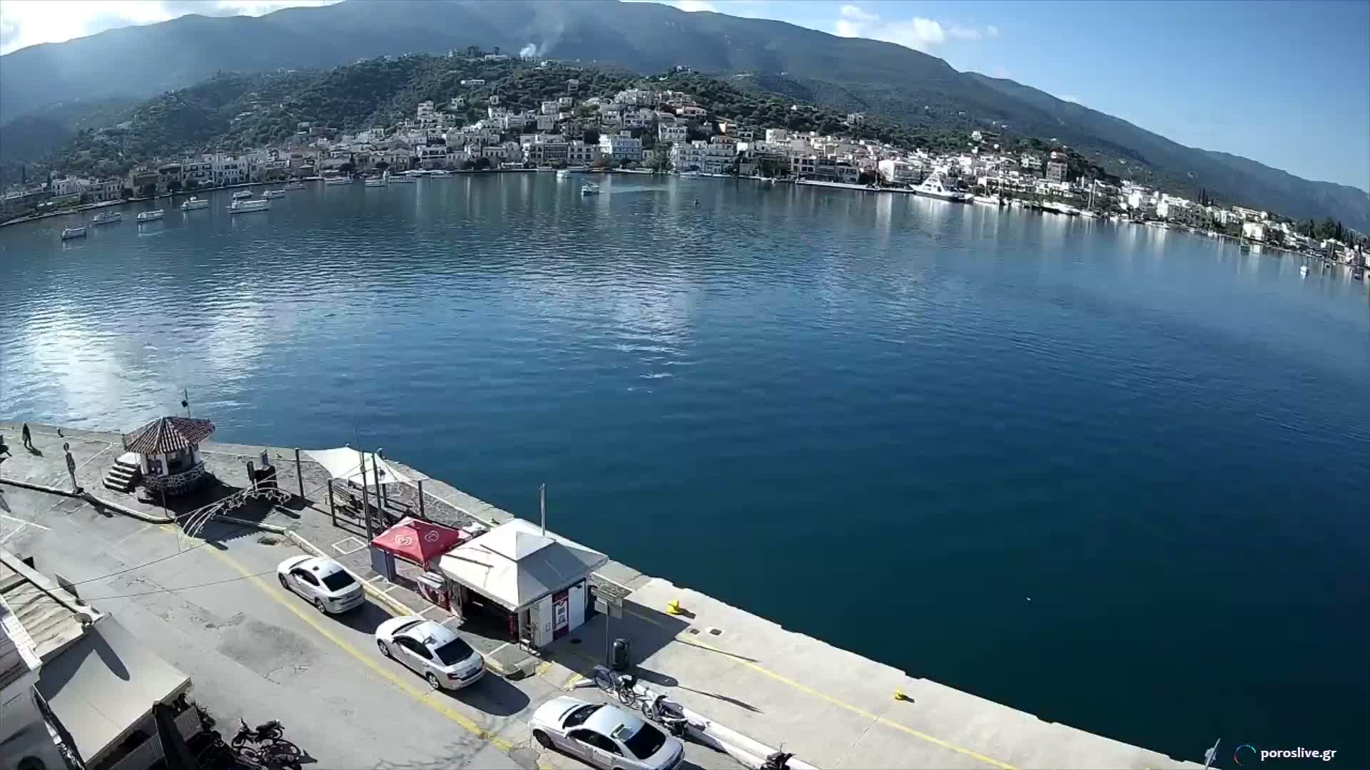 A bright, sunny day illuminates a vibrant blue bay, flanked by a bustling pier with cars in the foreground and a charming town of white buildings cascading up a green, mountainous hillside in the background.