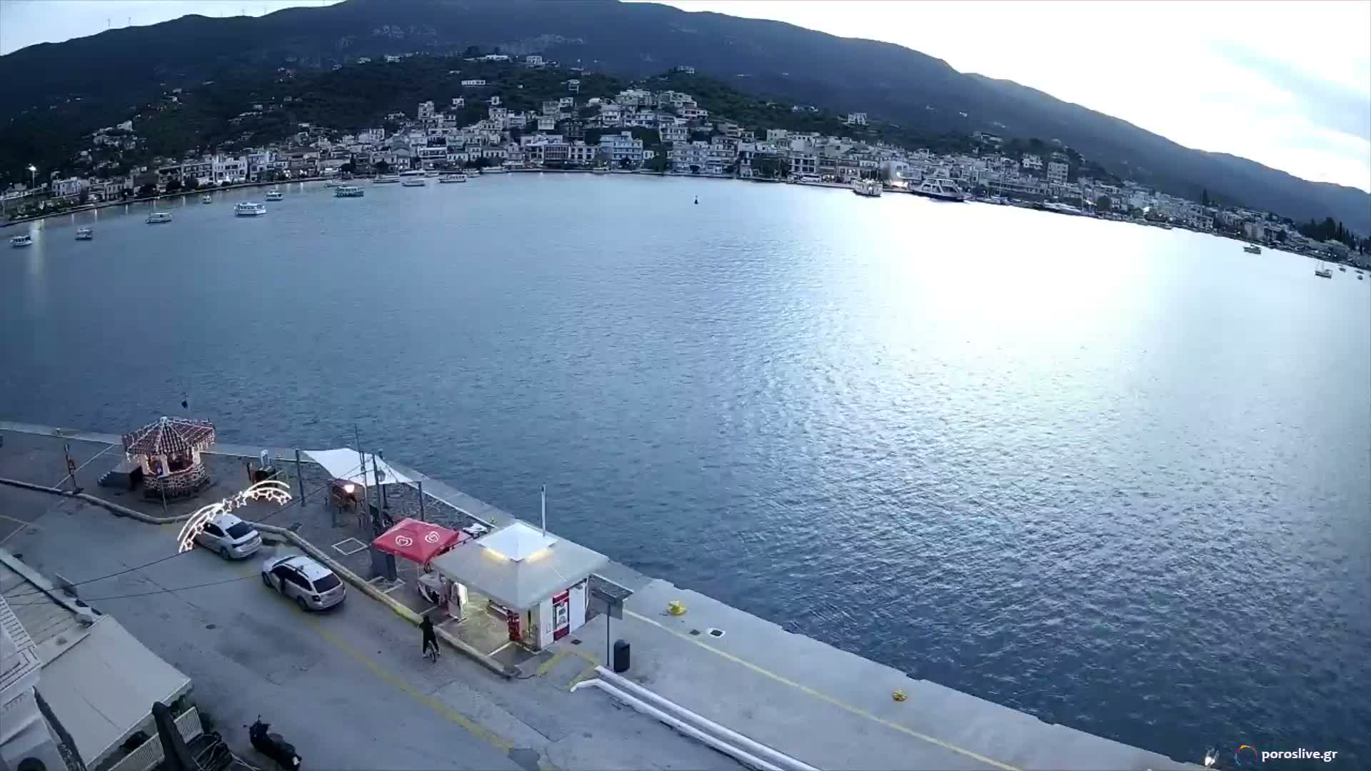 A bright, sunny day illuminates a vibrant blue bay, flanked by a bustling pier with cars in the foreground and a charming town of white buildings cascading up a green, mountainous hillside in the background.