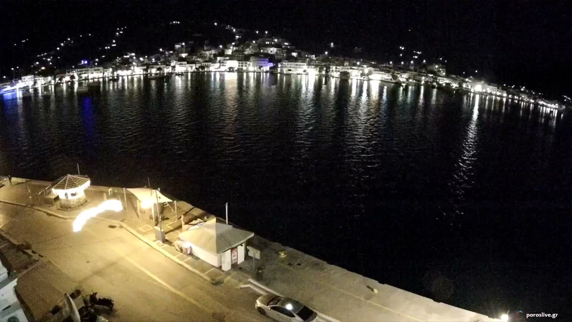 A clear night view shows a brightly lit waterfront town across calm dark waters, with a well-lit quay featuring a gazebo, various structures, and a parked car in the foreground.