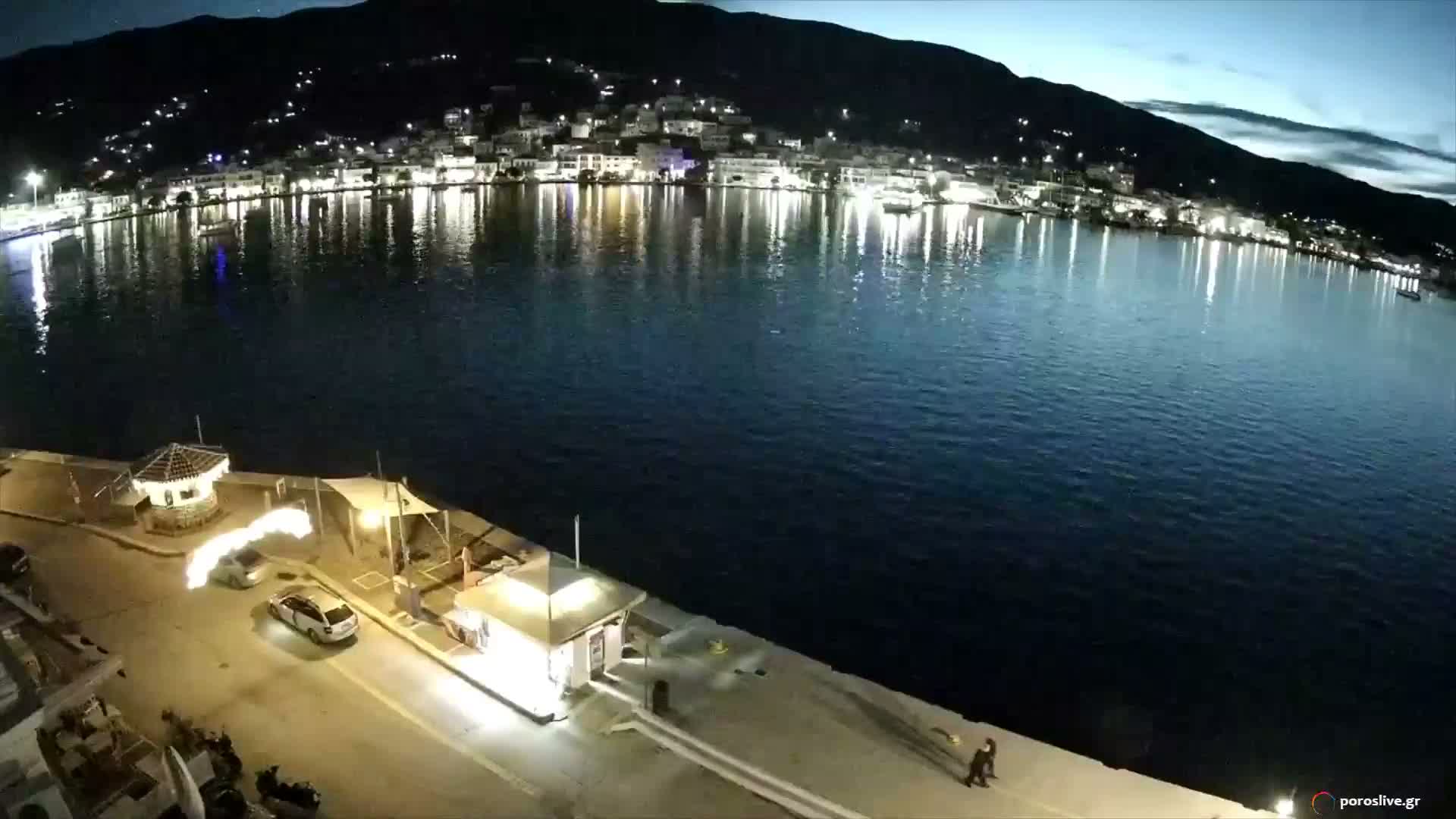 A well-lit pier with a few parked cars and pedestrians frames the foreground of a calm, dark harbor at night, with a brightly illuminated town reflecting on the water and distant mountains under a partly cloudy sky.