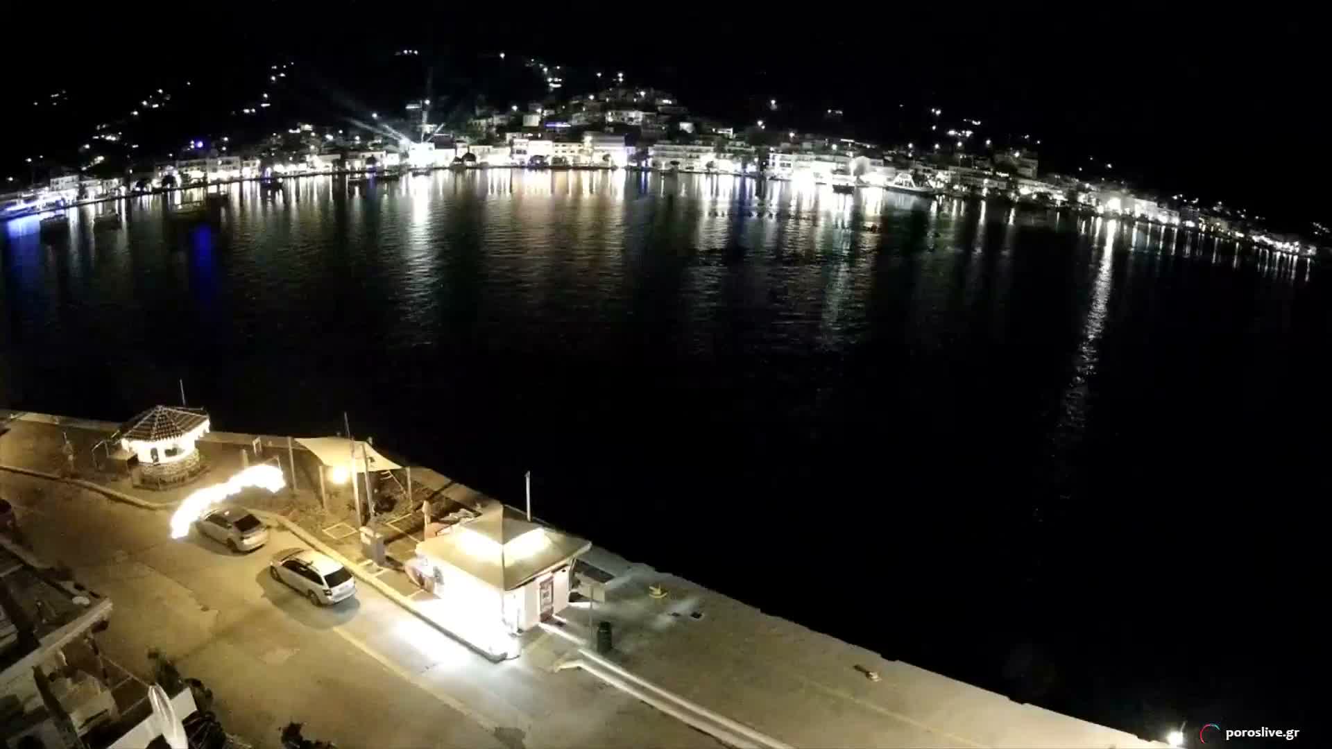 A well-lit pier with a few parked cars and pedestrians frames the foreground of a calm, dark harbor at night, with a brightly illuminated town reflecting on the water and distant mountains under a partly cloudy sky.