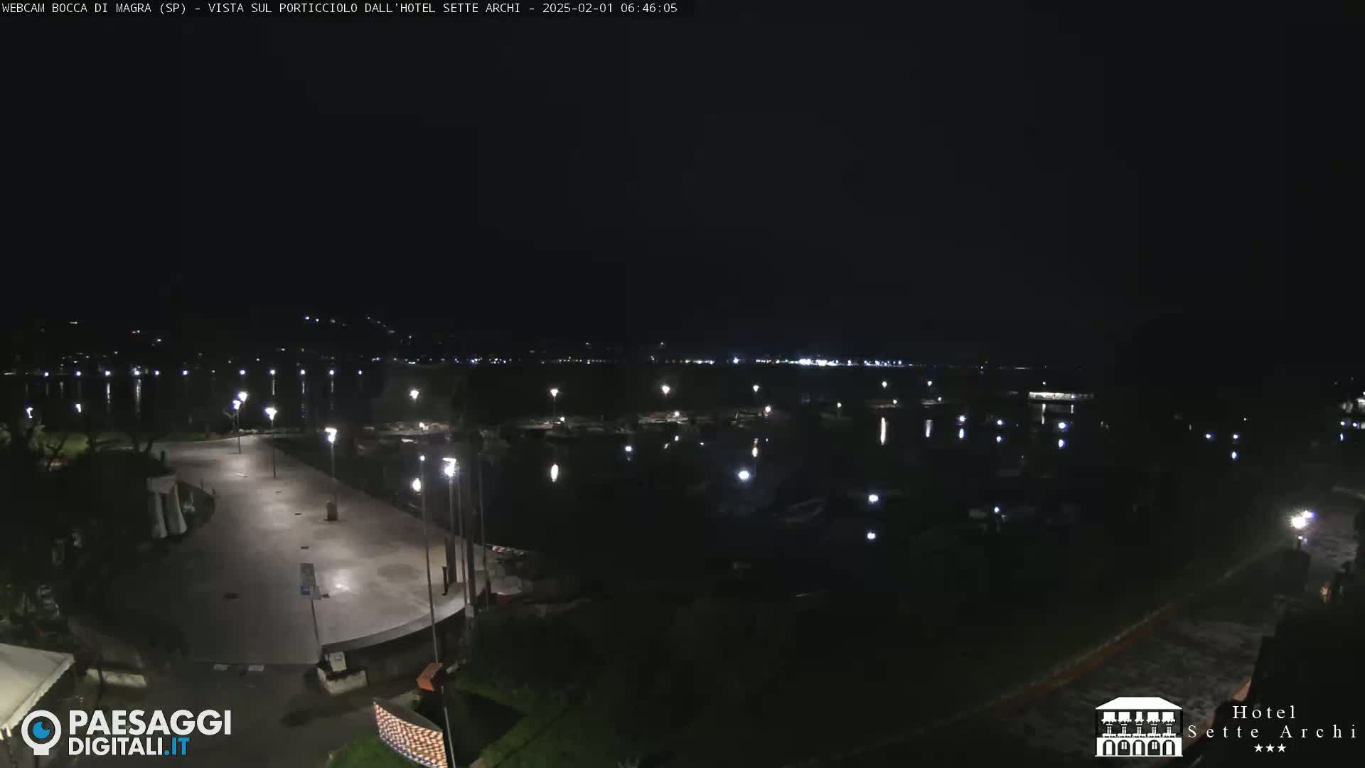 A nighttime view of a dark, calm body of water with a lit-up paved waterfront area and several boats.