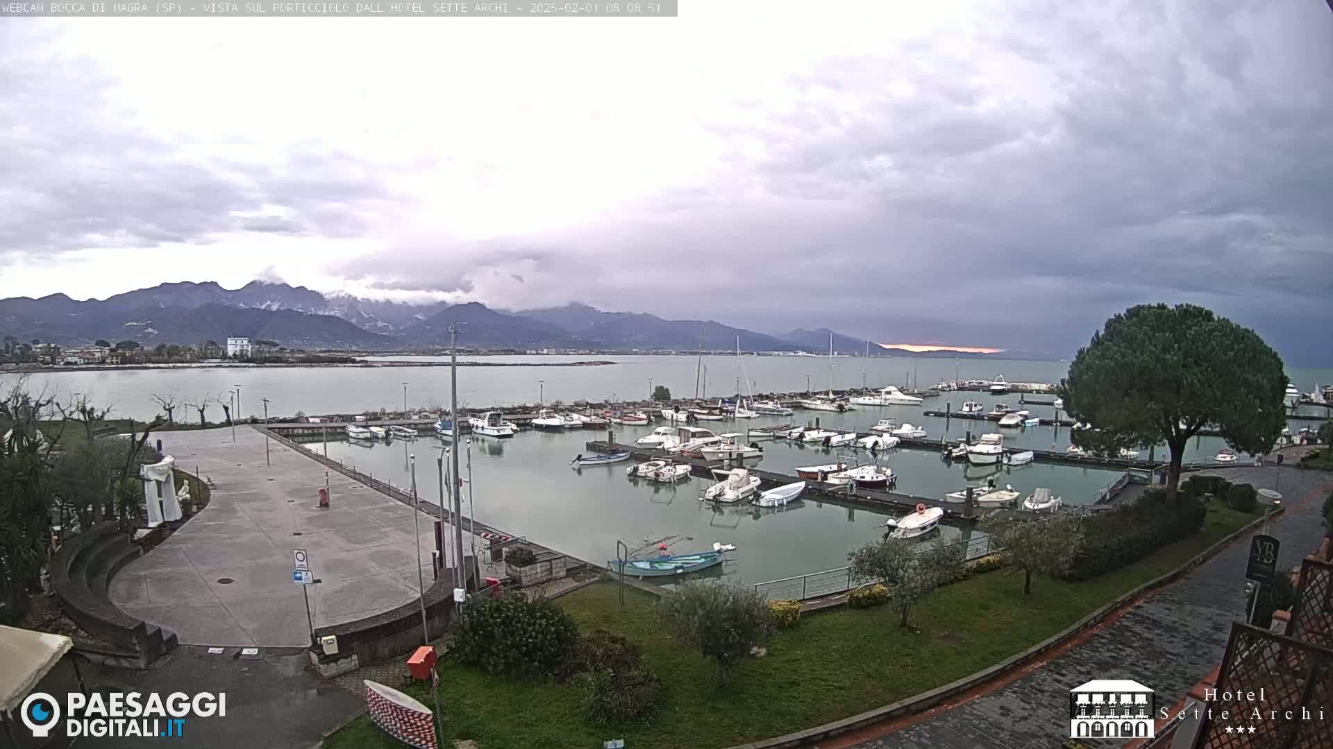 A marina filled with boats sits beside a paved plaza, with mountains in the distance under an overcast sky.