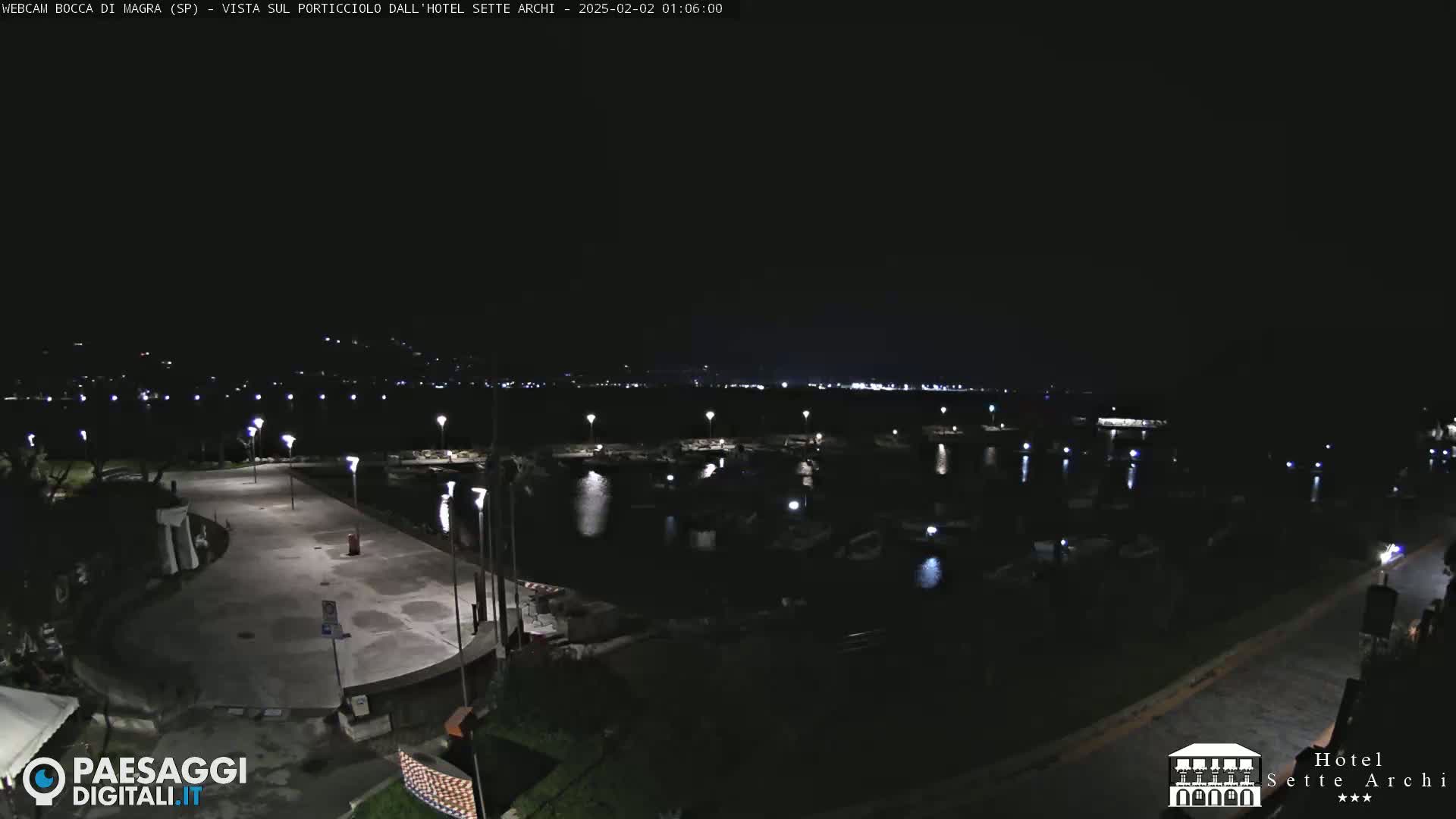 A nighttime view of a harbor filled with boats, a paved waterfront area, and dimly lit buildings in the distance.