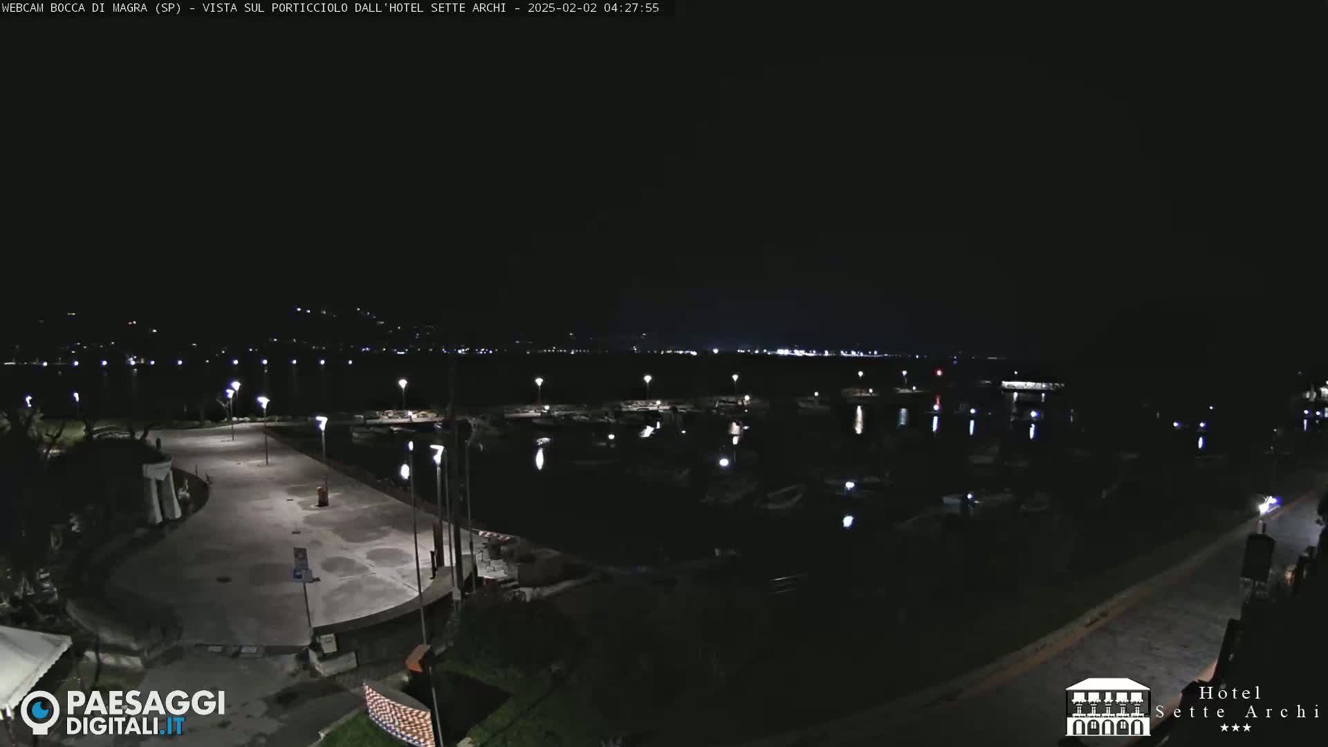 A nighttime view of a marina with boats docked, illuminated by lights along a paved waterfront area.