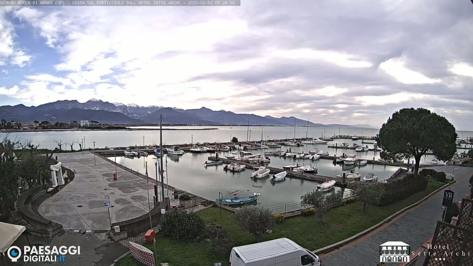 A harbor filled with numerous boats is shown under a cloudy sky, with snow-capped mountains visible in the distance.