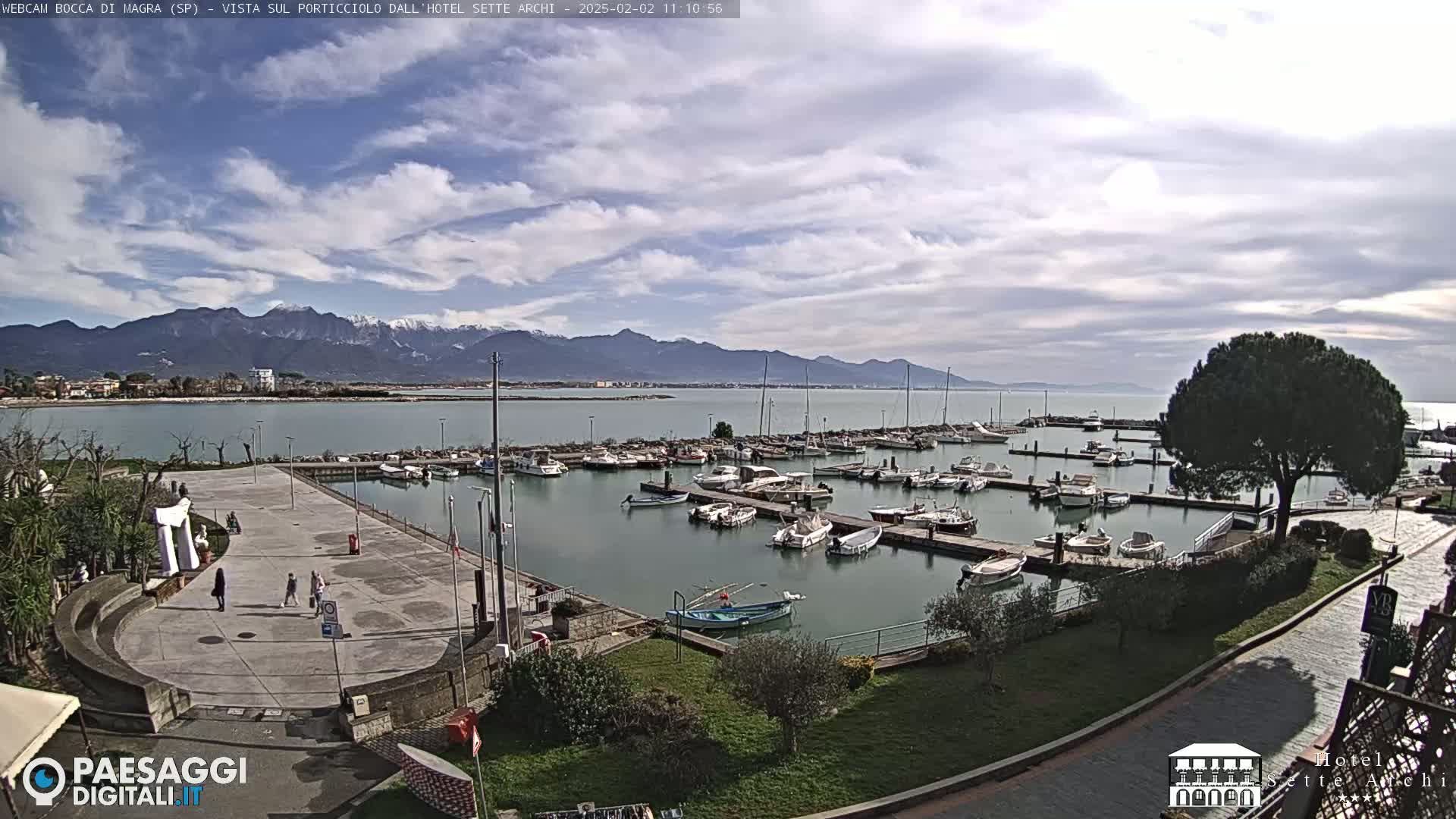 A partly cloudy day overlooks a marina filled with boats, a paved plaza, and snow-capped mountains in the distance.