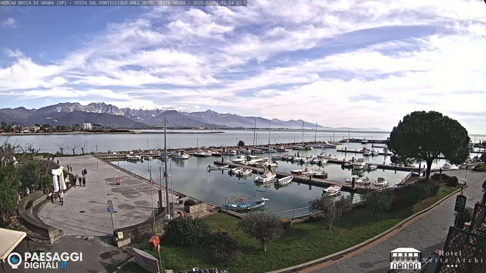 A harbor filled with boats is situated below a partly cloudy sky and in front of snow-capped mountains.