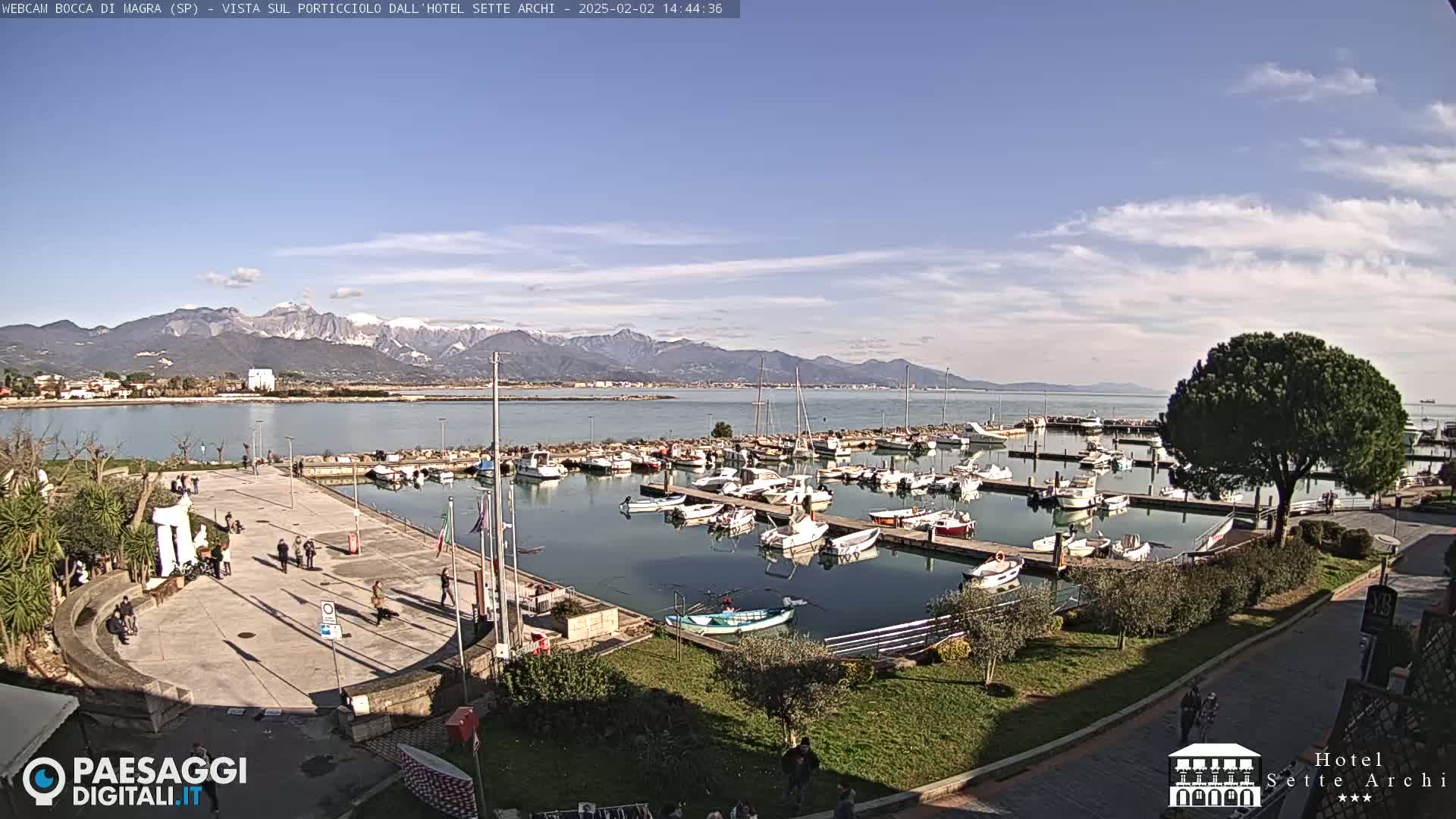 A sunny day shows a harbor filled with boats, a paved area with people walking, and snow-capped mountains in the distance.