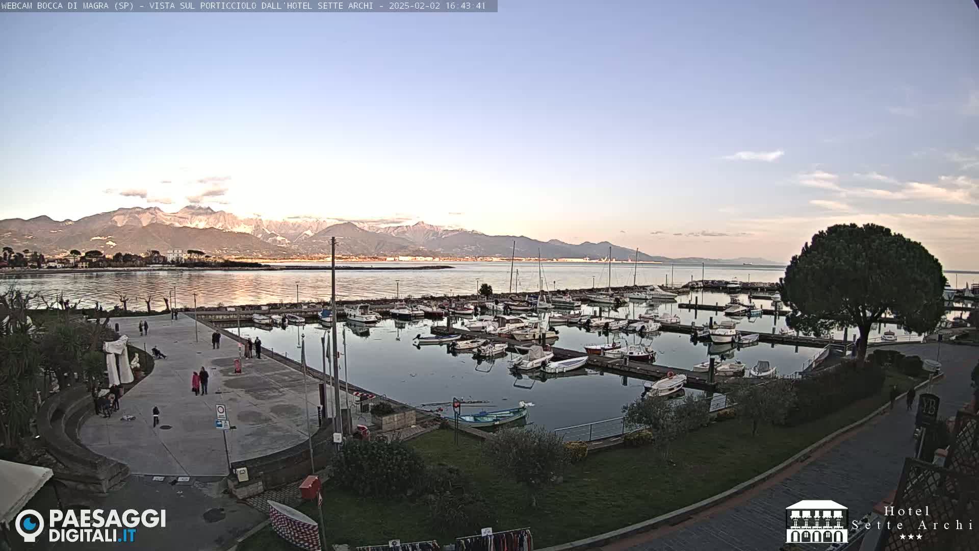 A calm harbor filled with docked boats is situated near a paved plaza with a few people walking, overlooking a body of water and mountains under a clear, twilight sky.