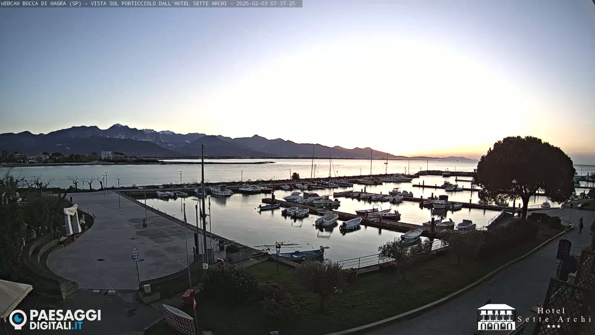 A calm harbor filled with numerous boats is nestled against a shoreline, with mountains in the background under a twilight sky.