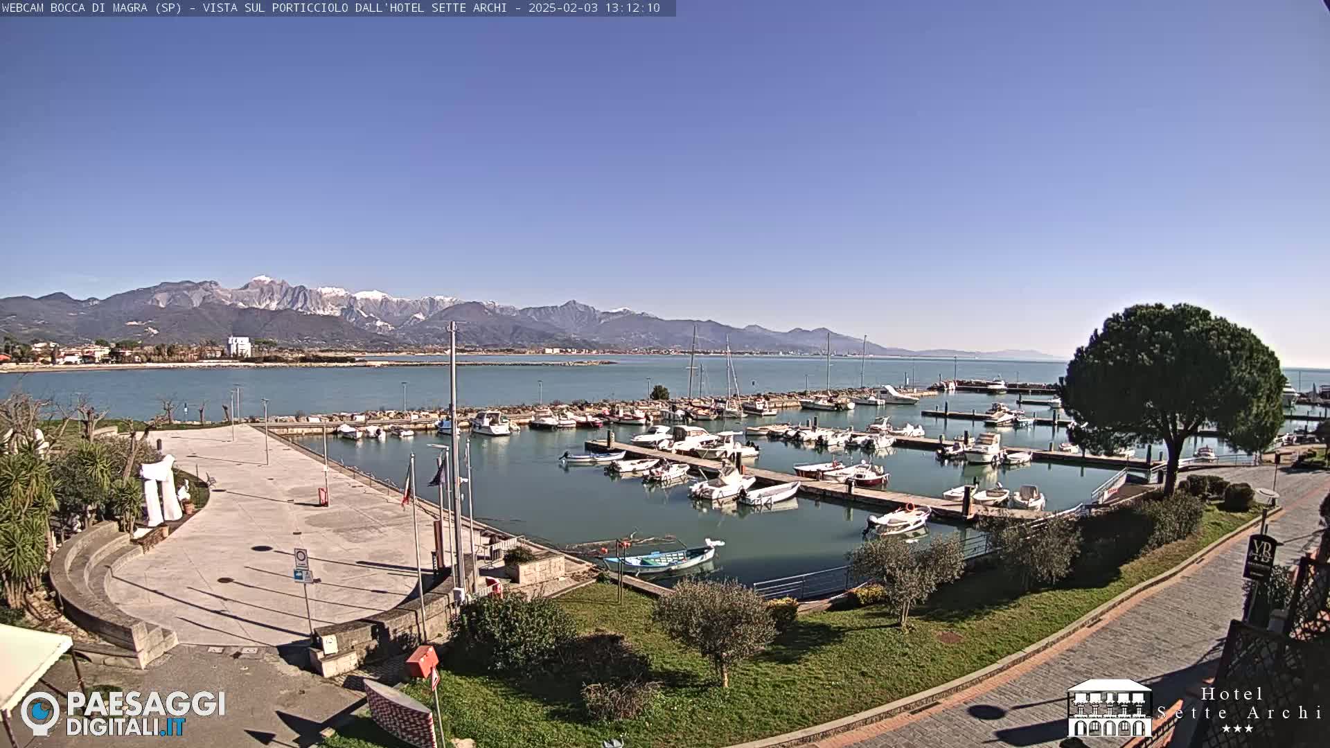 A marina filled with boats sits beside a paved plaza, overlooking calm water and snow-capped mountains under a clear blue sky.