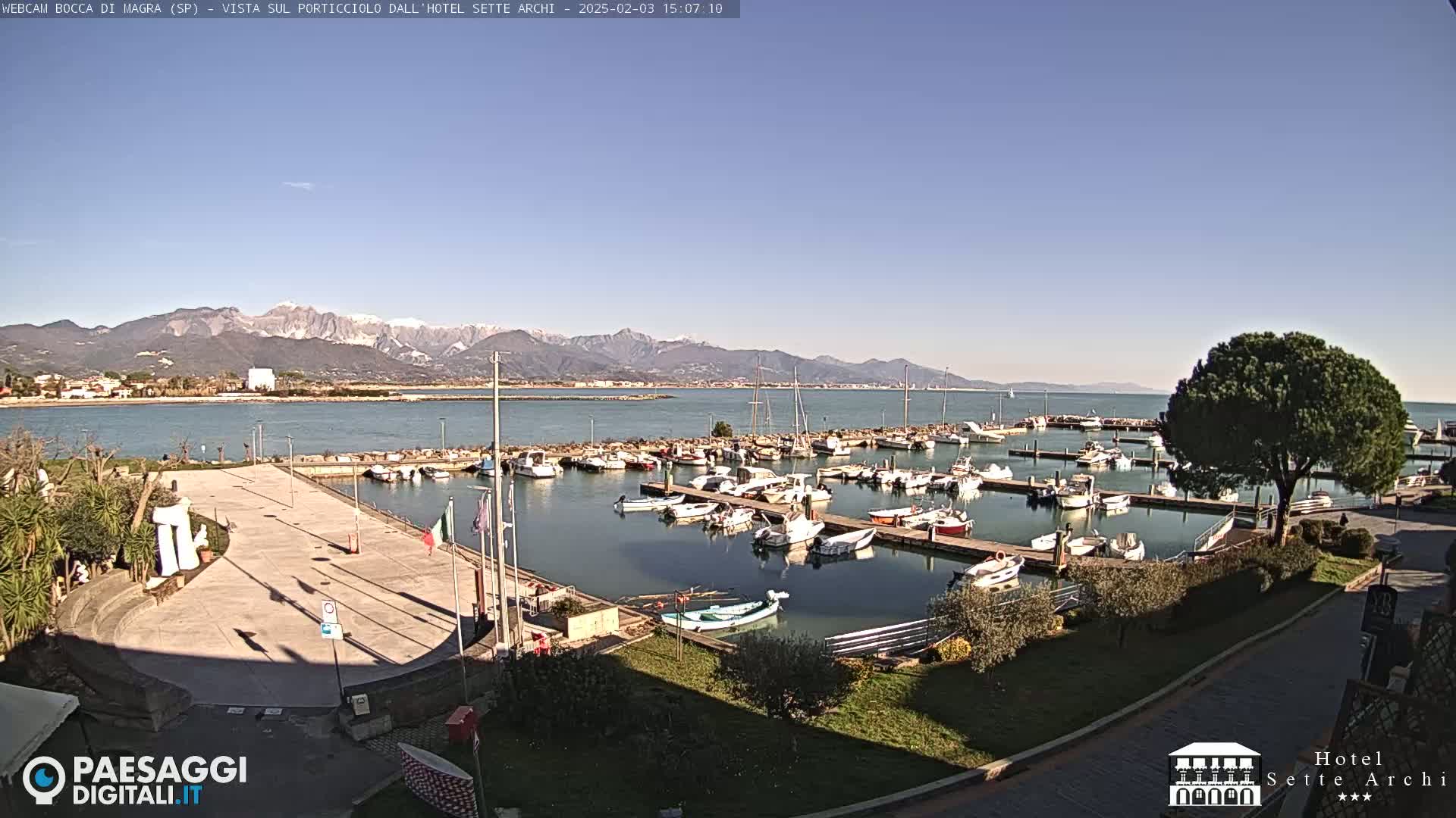 A harbor filled with numerous boats is situated near a plaza, with snow-capped mountains visible in the distance under a clear sky.
