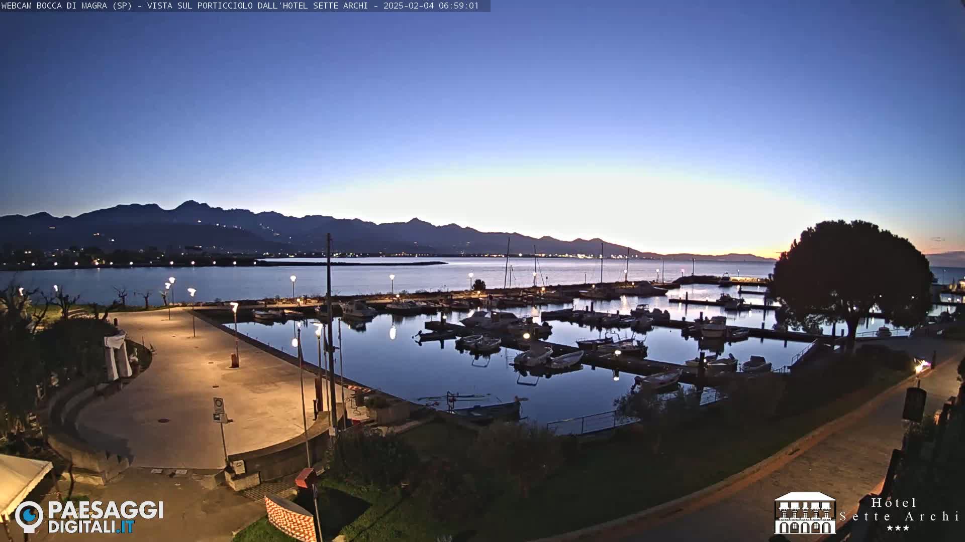 A marina filled with boats at twilight, situated next to a paved walkway and overlooking a calm body of water with mountains in the distance.