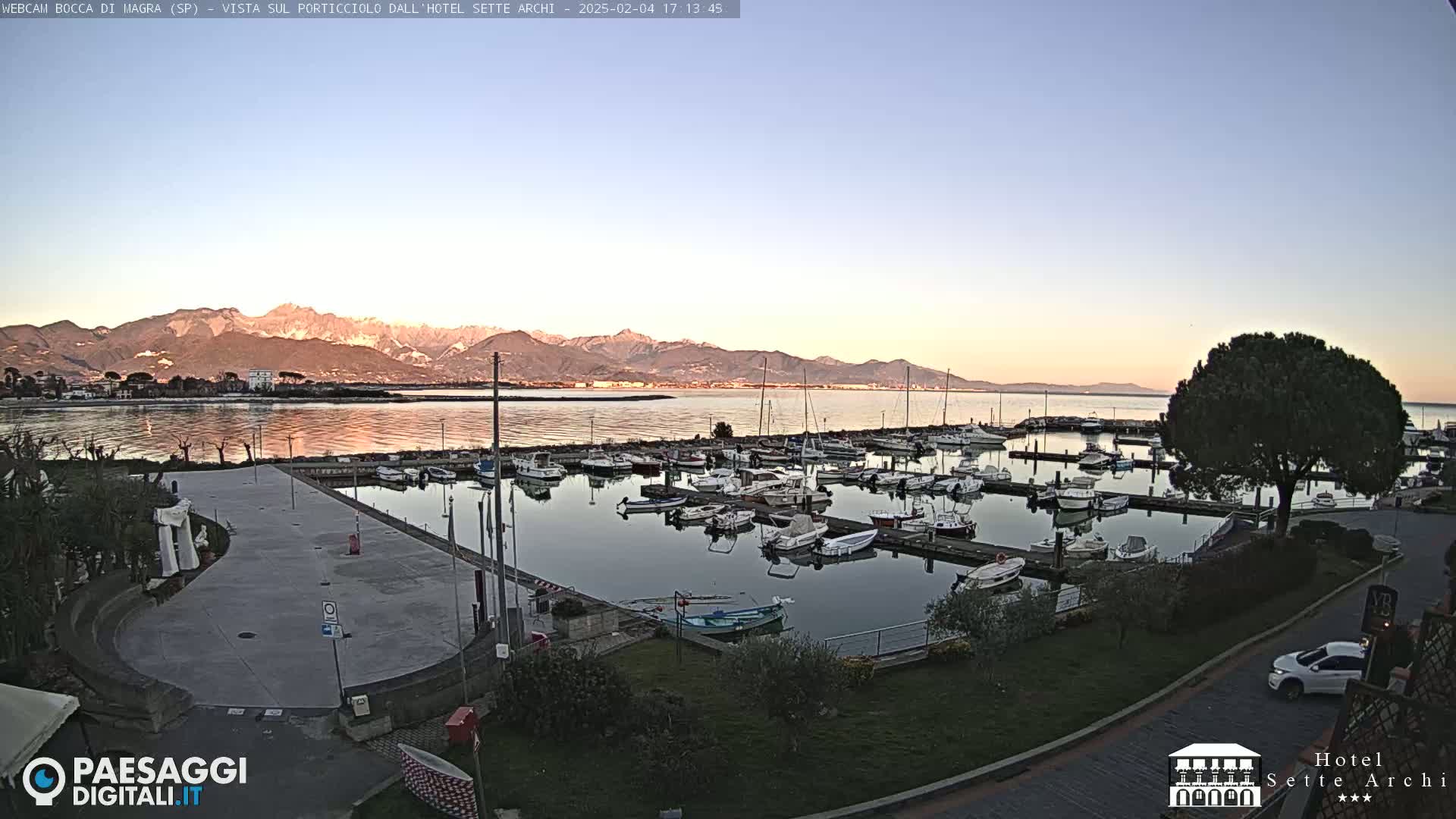 A calm marina filled with numerous boats is situated near a waterfront plaza, with mountains visible in the distance under a clear, twilight sky.