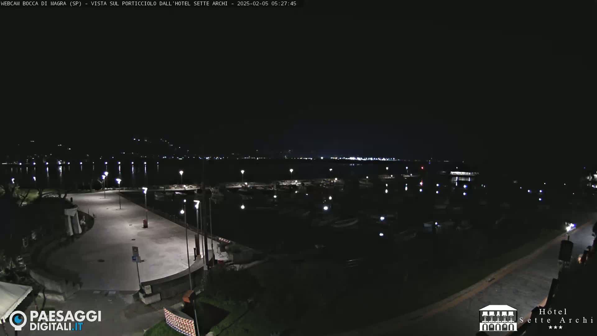 A nighttime view of a harbor filled with boats, a paved waterfront area, and distant lights across the water.