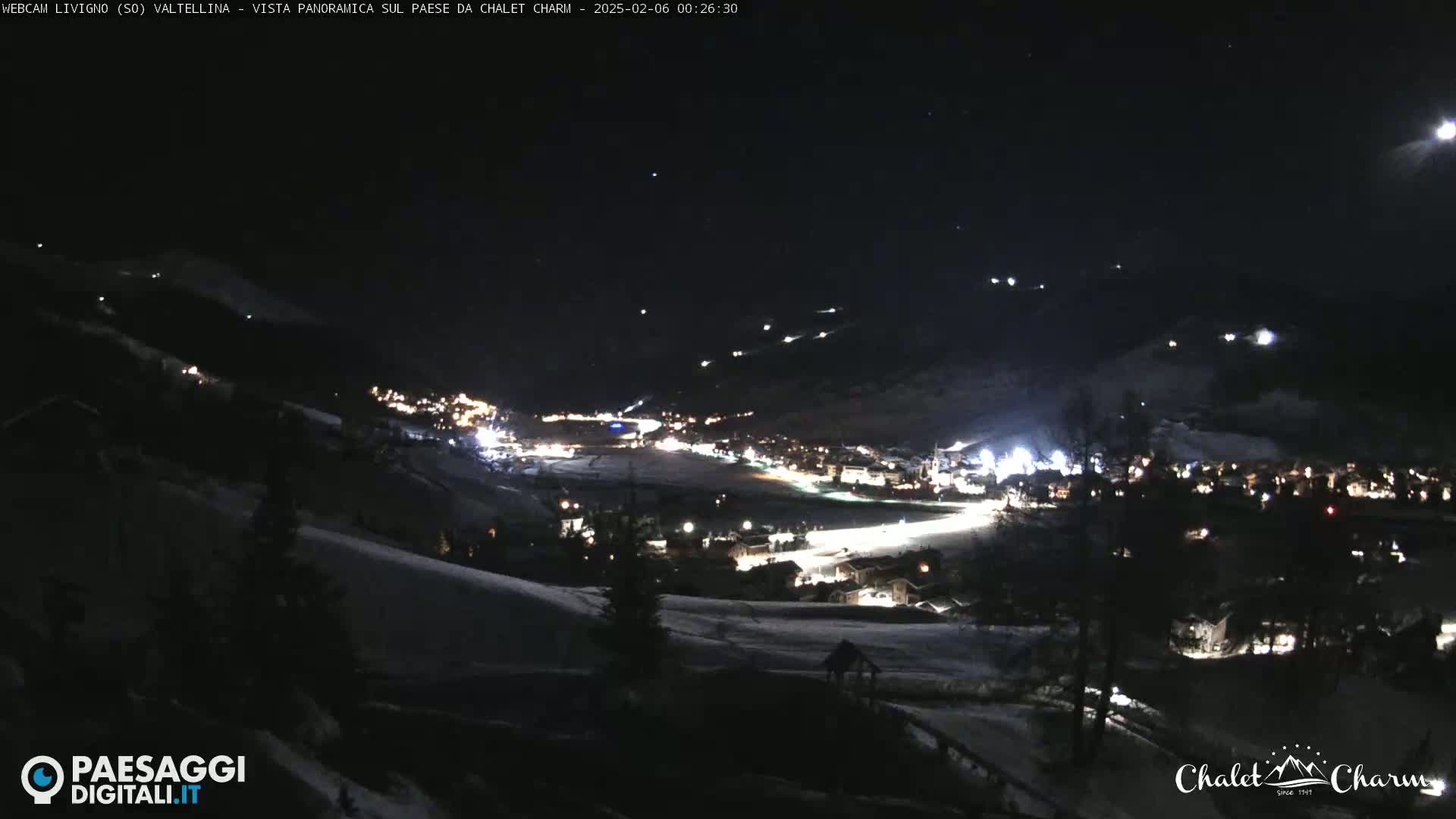 A nighttime, snow-covered mountain town is illuminated by lights, with a moon visible in the dark sky.