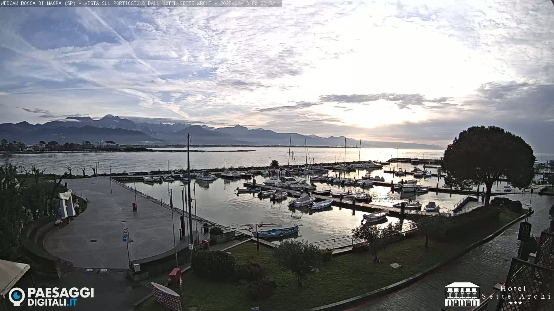 A marina filled with boats sits next to a paved plaza, all overlooking calm water with distant mountains under a cloudy sky.