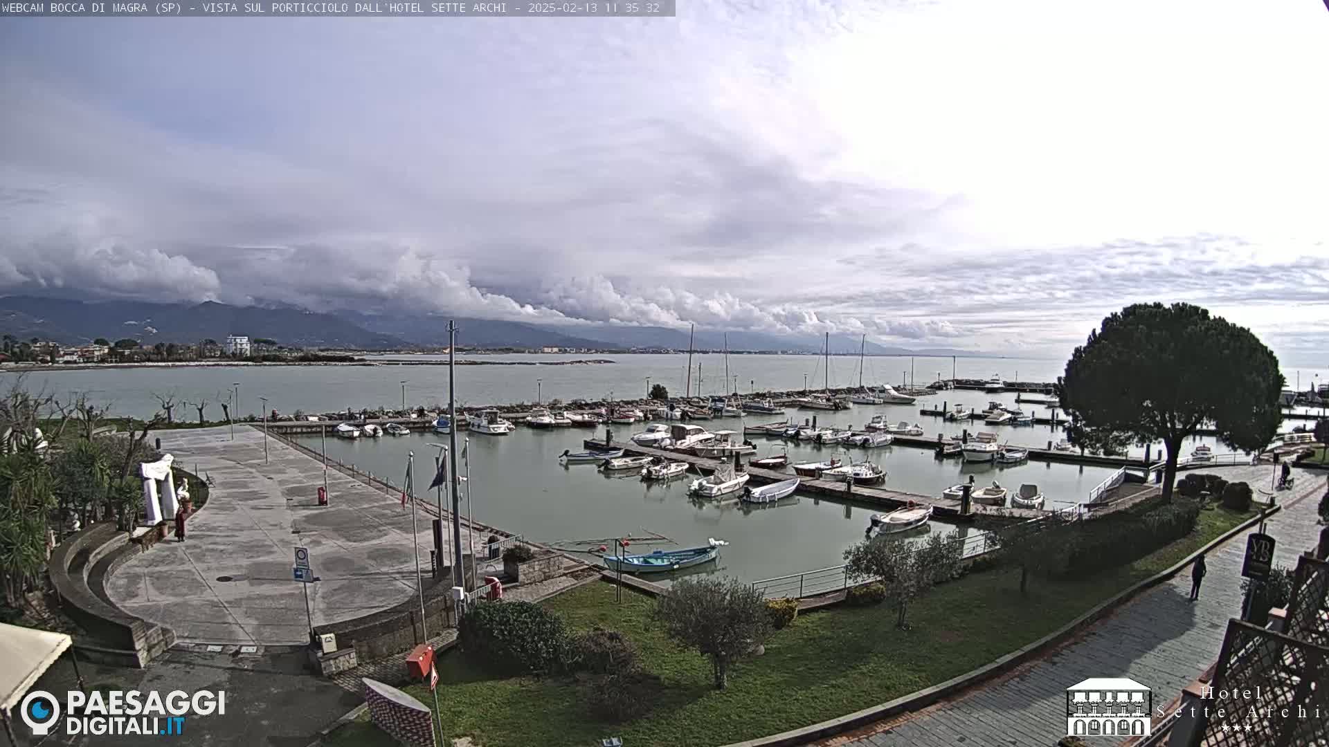 A marina filled with boats sits on calm water under a cloudy sky, with mountains visible in the distance.
