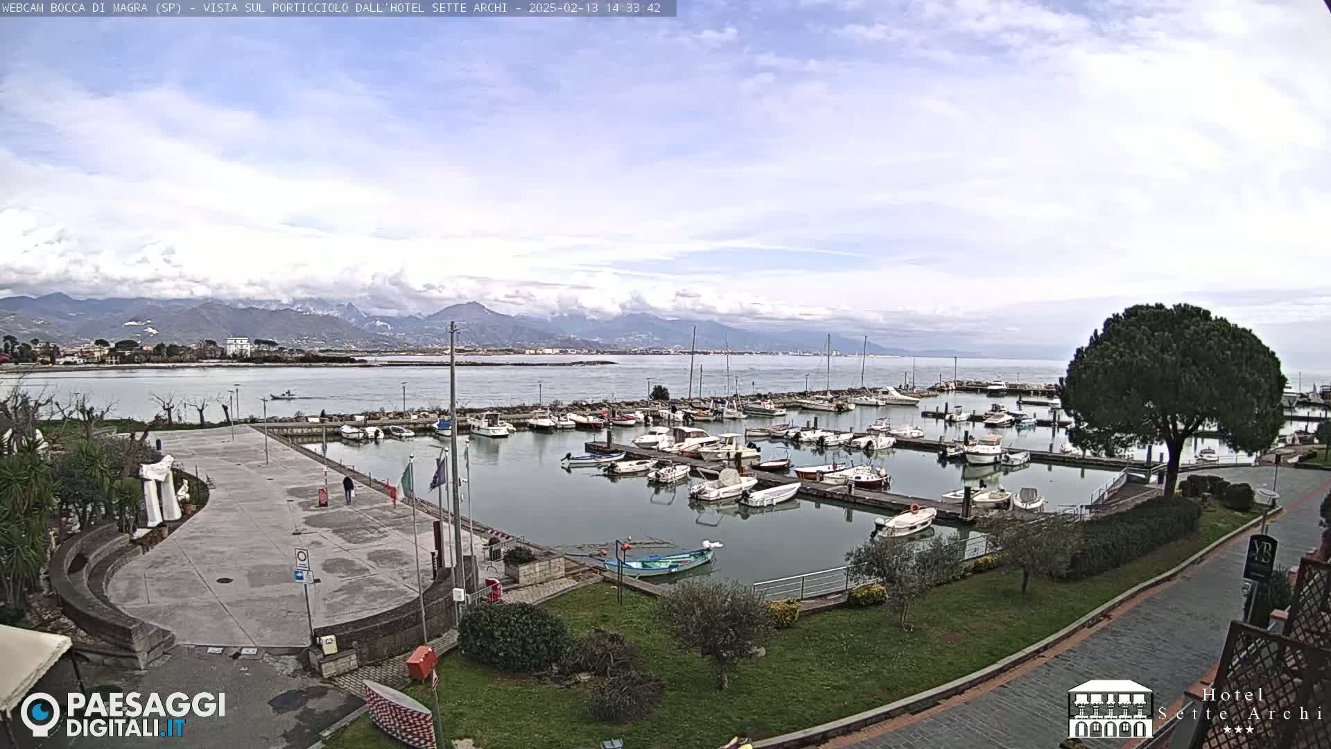 A marina filled with boats is situated near a paved plaza and overlooks a calm body of water with mountains in the background under a partly cloudy sky.