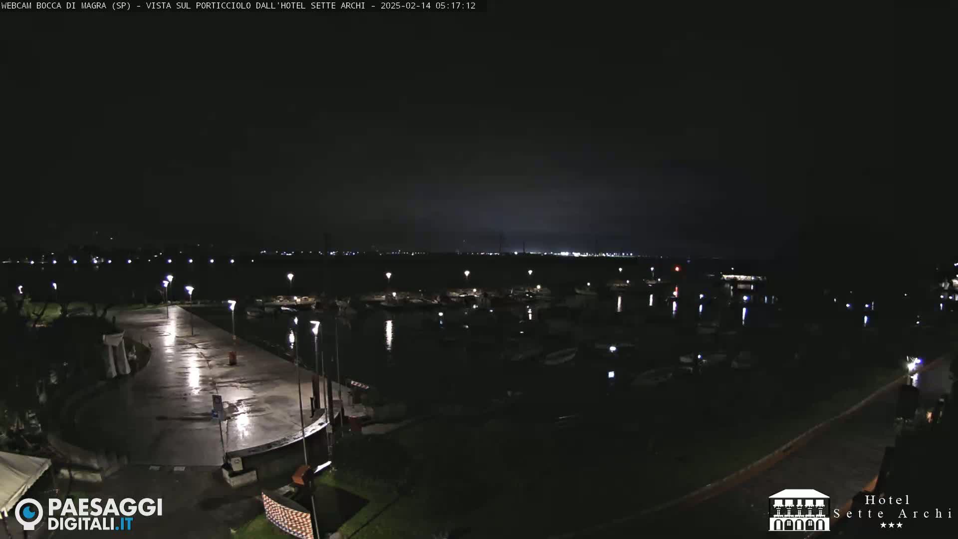 A nighttime view of a wet, empty paved area next to a marina filled with boats under a dark sky.